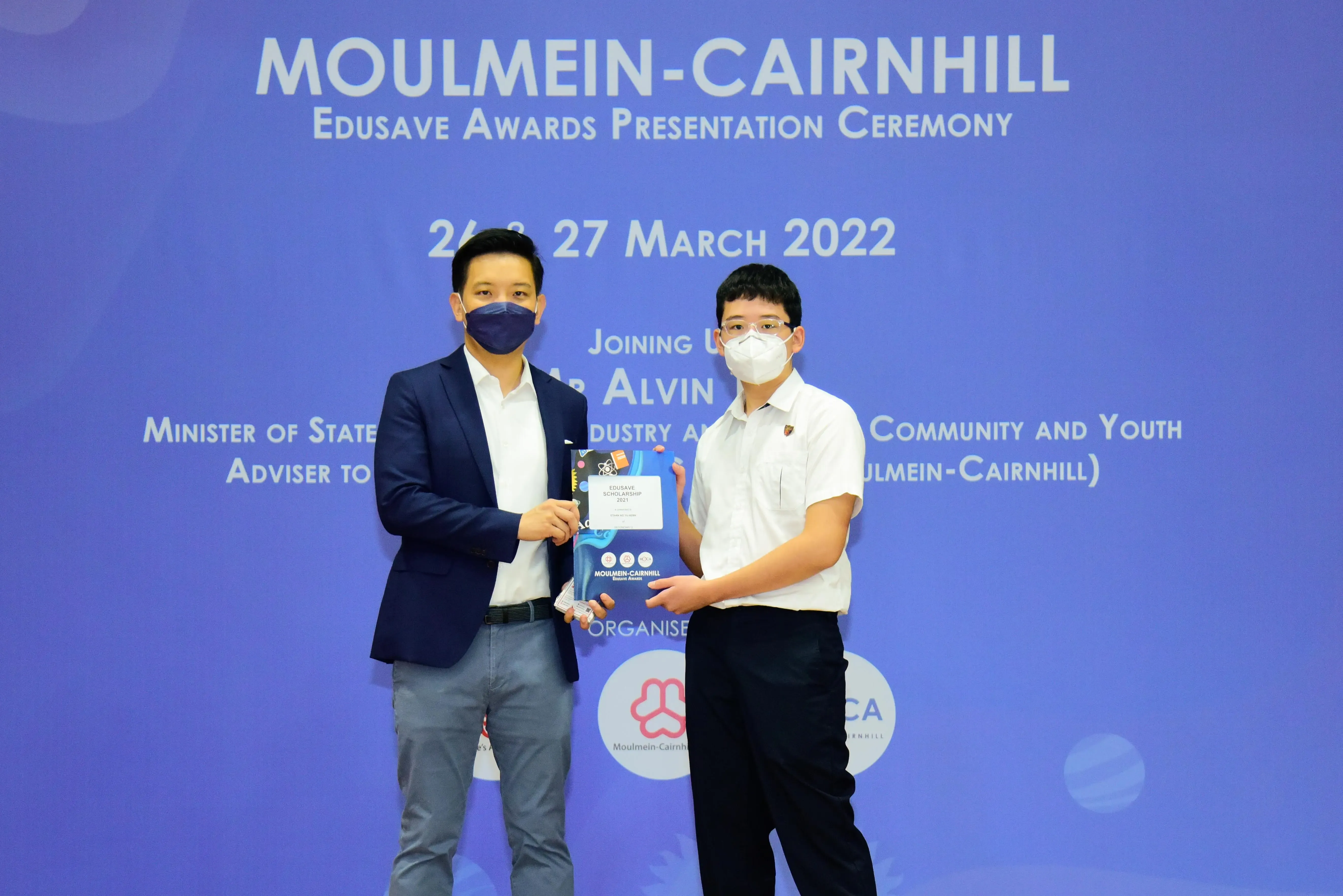 Two masked individuals hold an Edusave Scholarship certificate at the Moulmein-Cairnhill Awards Ceremony, March 2022.