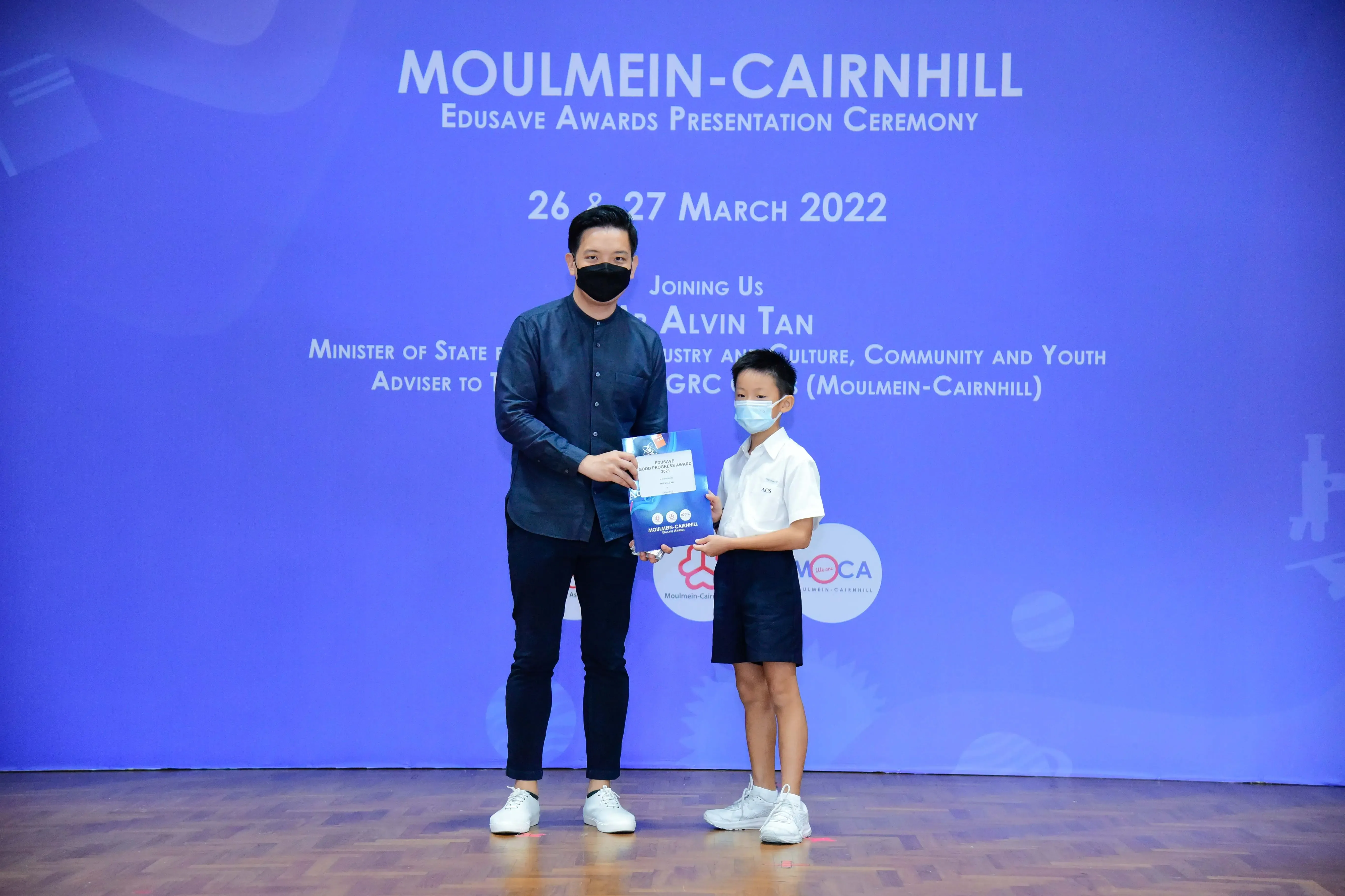 An adult and a young boy in masks hold a certificate at the Moulmein-Cairnhill Edusave Awards Ceremony, March 2022.