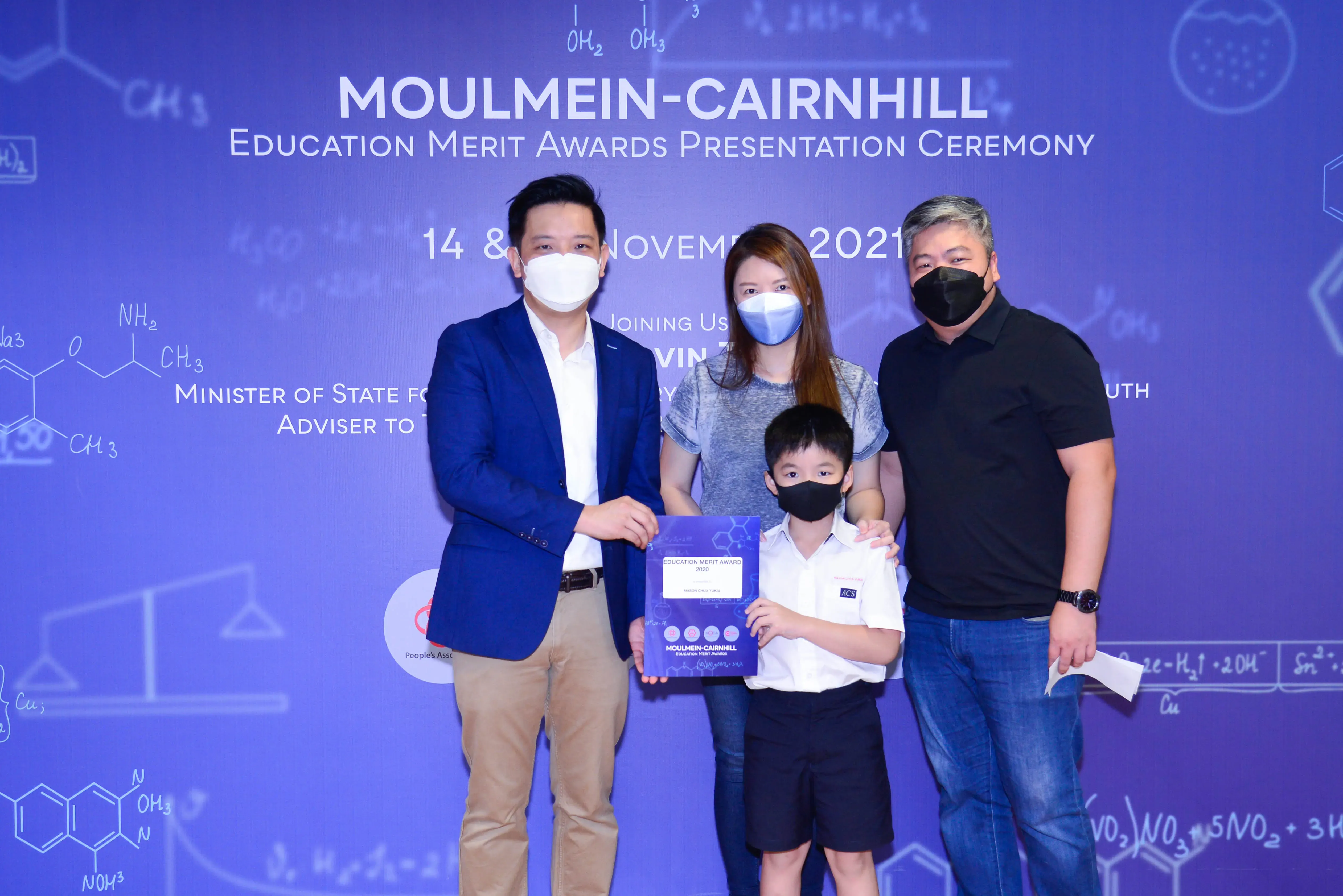 Four masked people pose at the Moulmein-Cairnhill Education Merit Awards Ceremony, with a boy holding an award plaque.