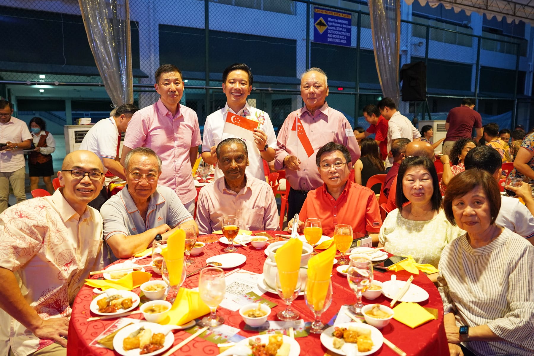 Group of people posing at a red-clothed dinner table outdoors, some holding small Singapore flags.