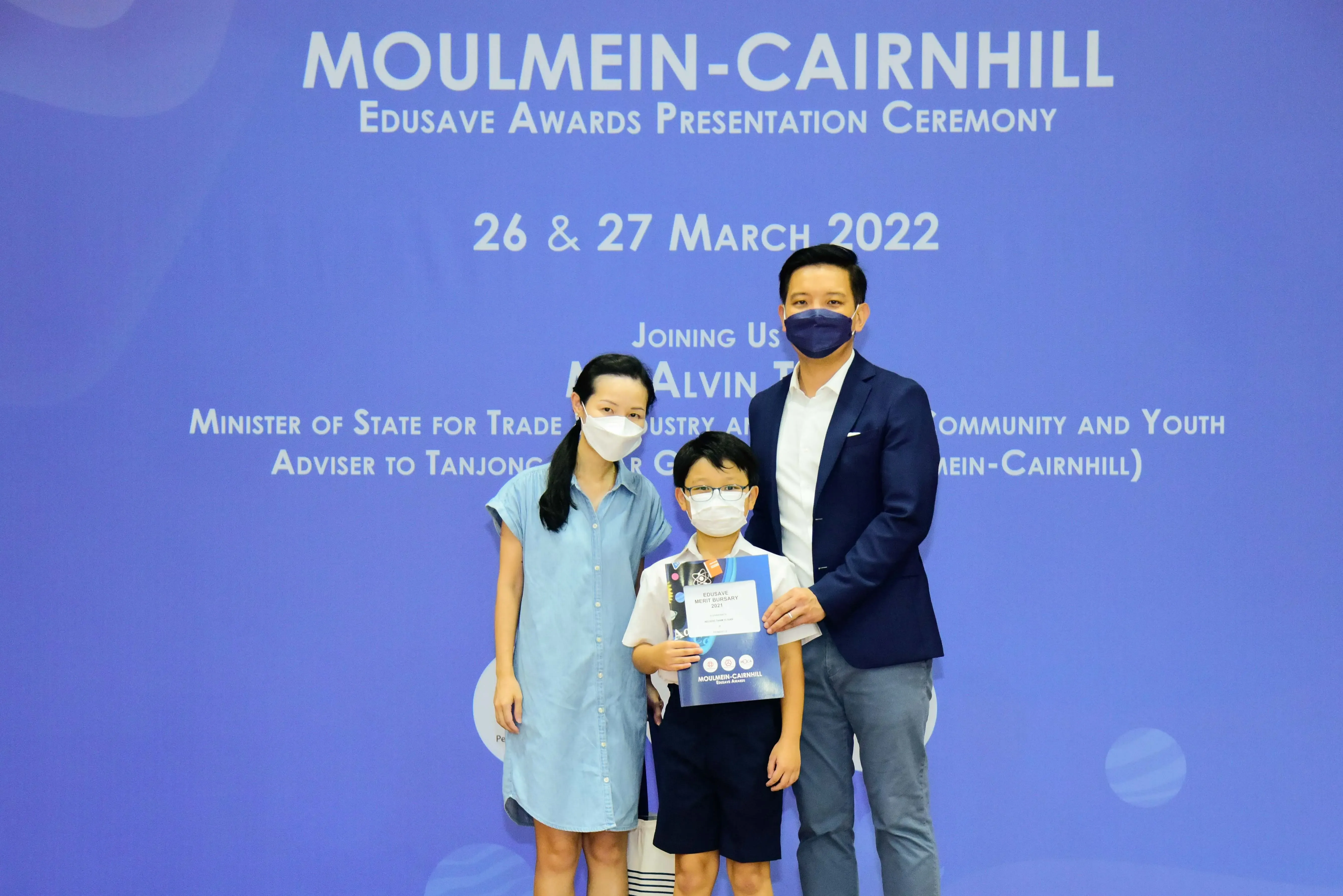 Three masked people pose before a Moulmein-Cairnhill Edusave Awards Presentation Ceremony banner dated March 2022.