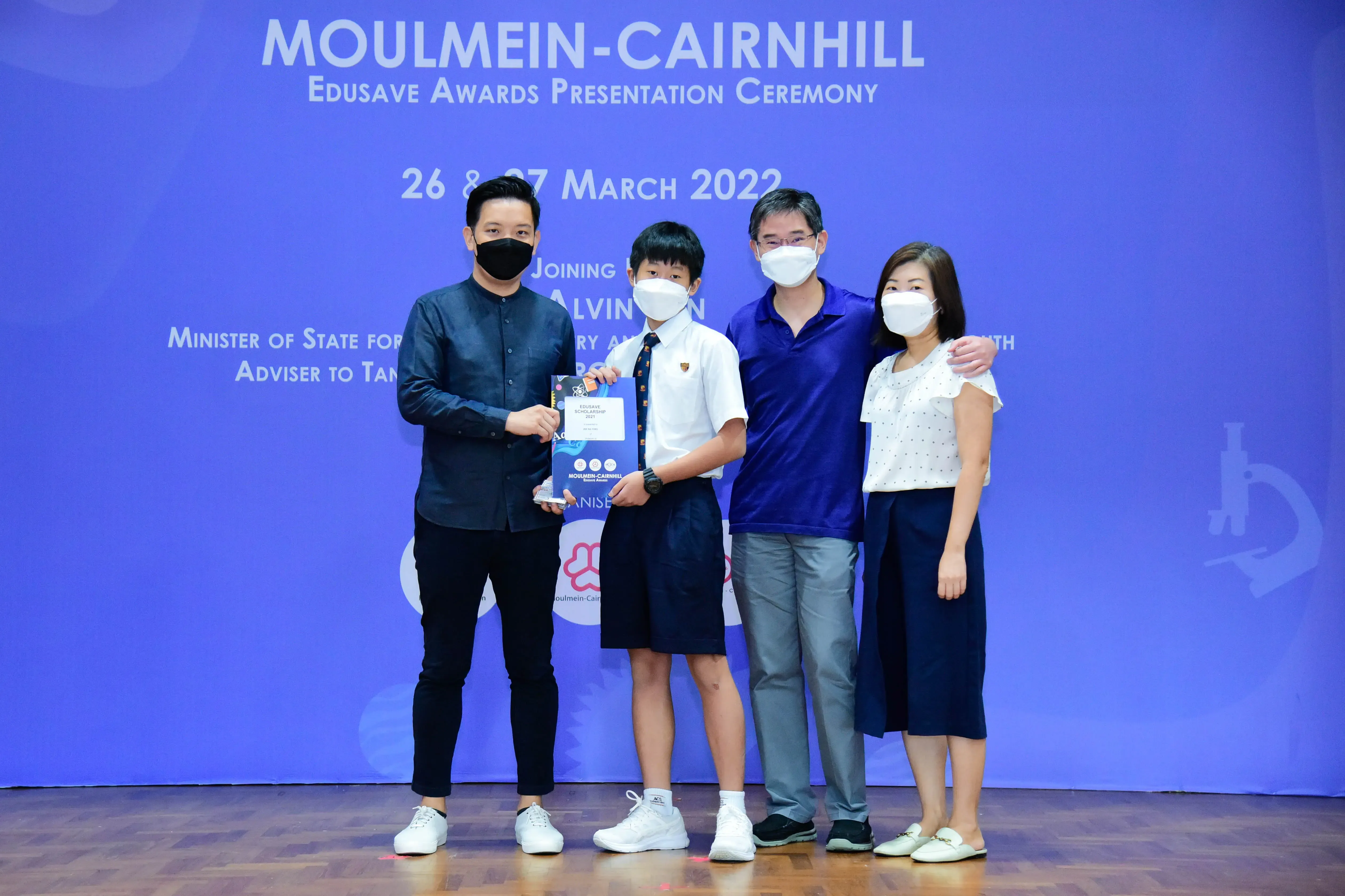 Four masked people pose at the Moulmein-Cairnhill Edusave Awards Ceremony, with a student holding a trophy.