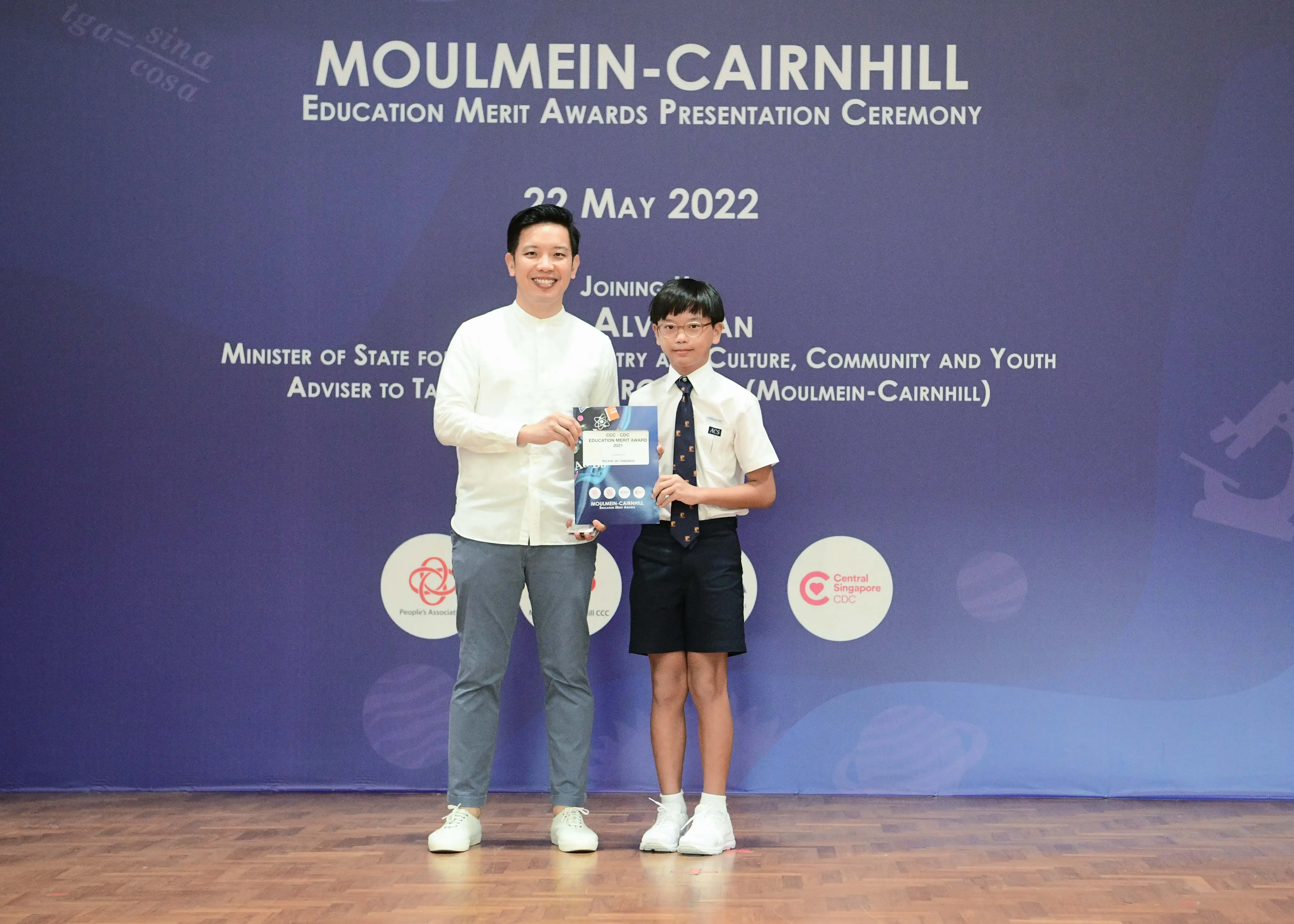 An adult and a young boy in school uniform hold an award certificate at the Moulmein-Cairnhill Education Merit Awards 2022.