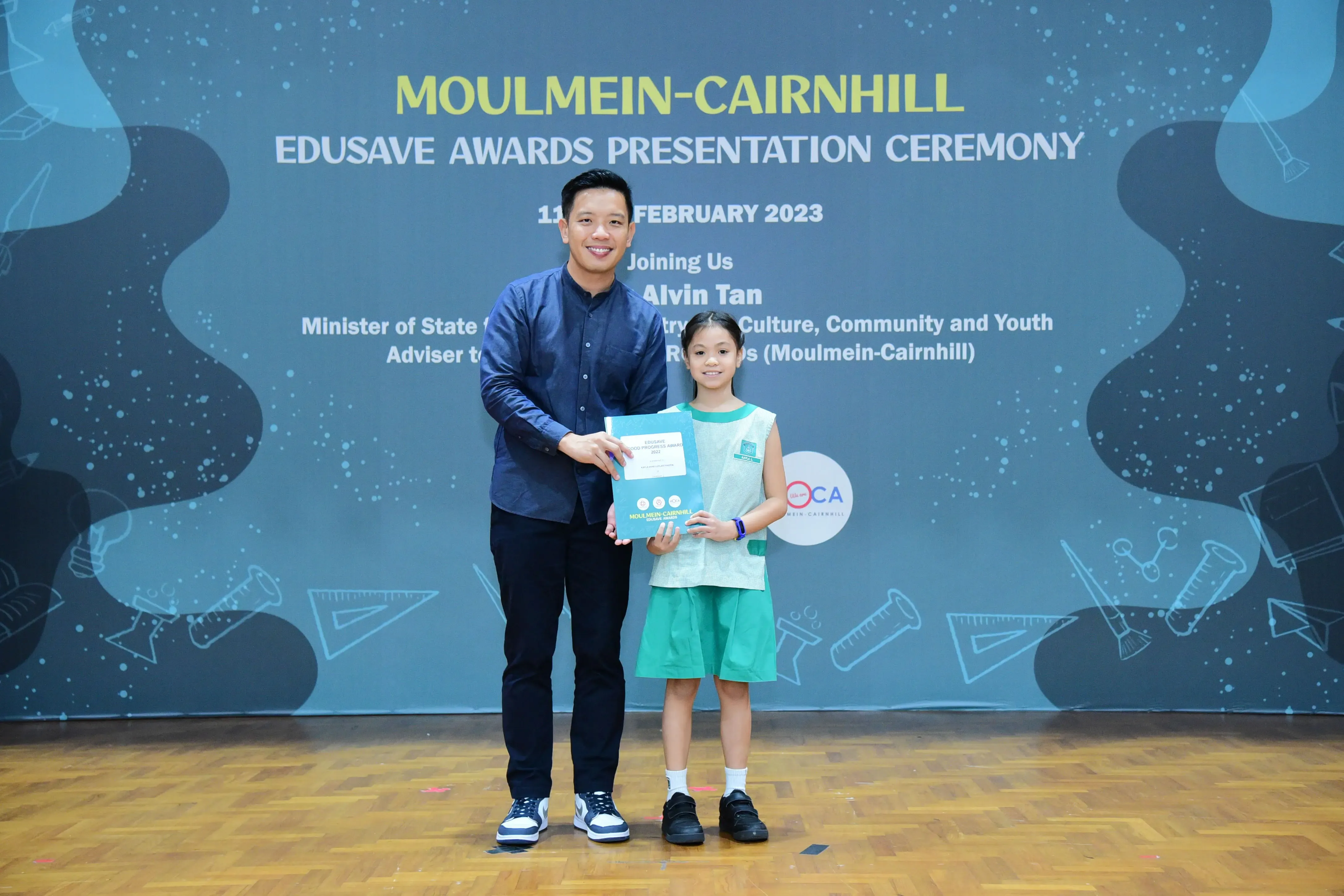 A man and a young girl in school uniform hold an award together at the Moulmein-Cairnhill Edusave Awards Ceremony 2023.