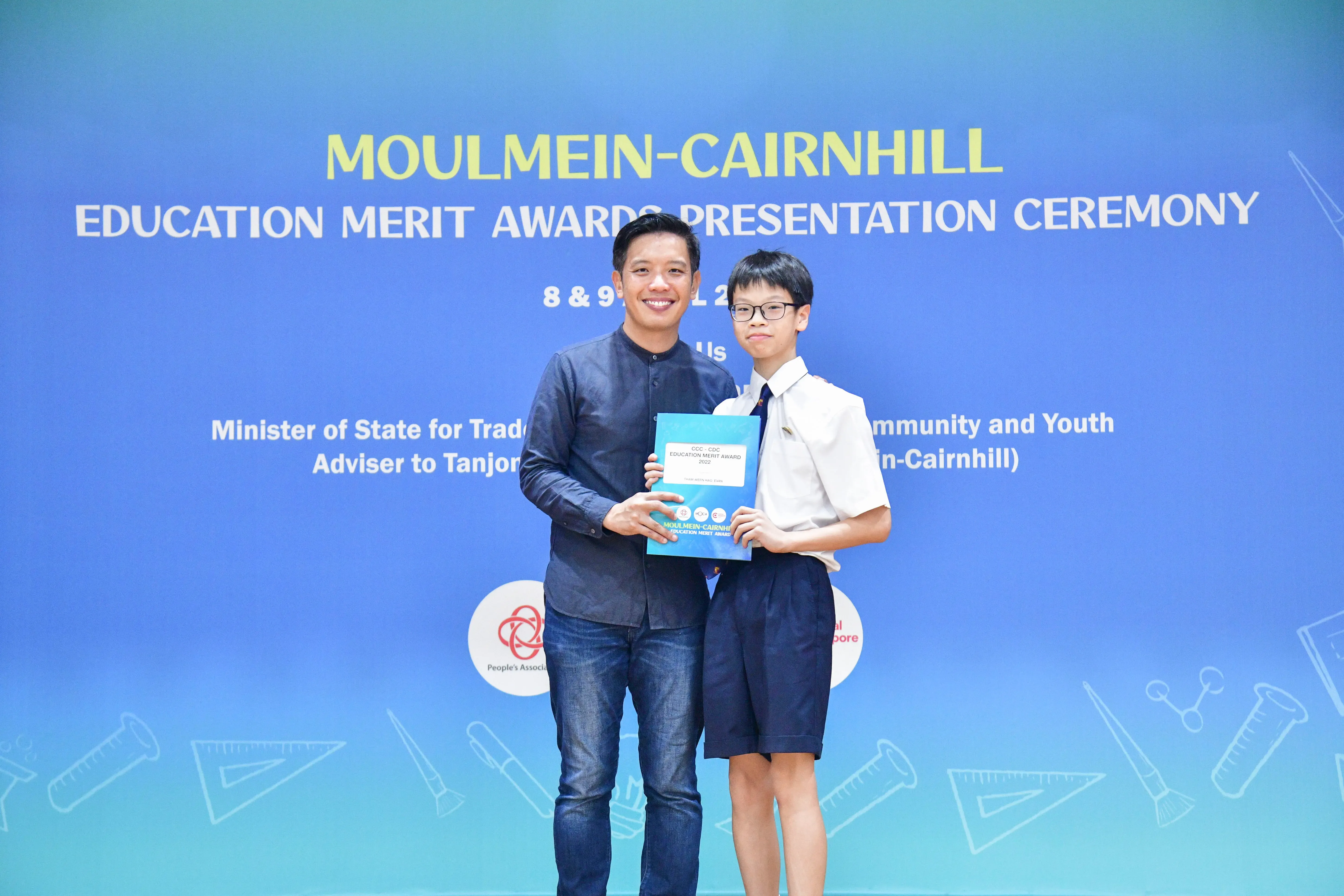 An adult and a young student in uniform hold a certificate at the Moulmein-Cairnhill Education Merit Awards ceremony.