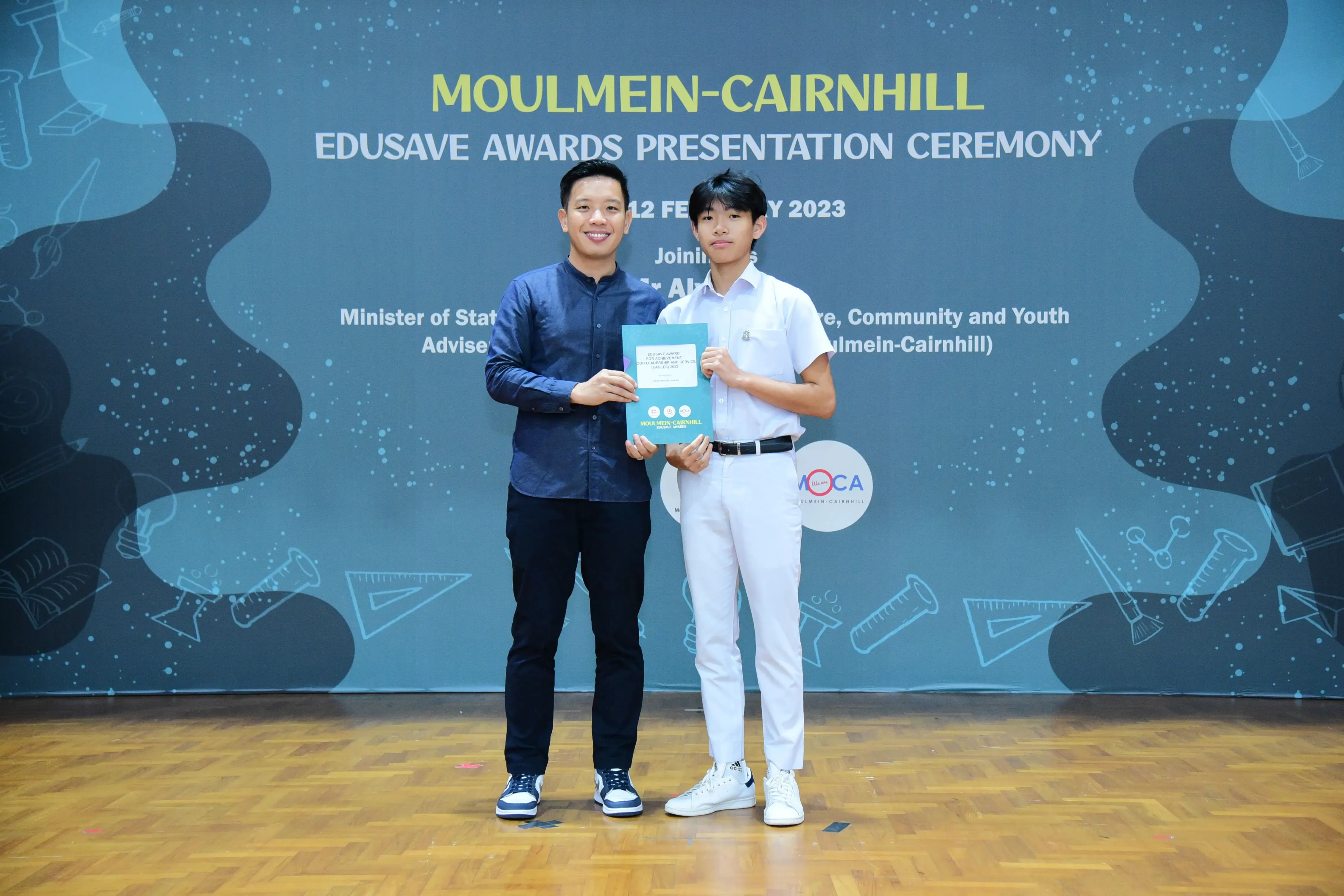 Two people hold a booklet together on stage at the Moulmein-Cairnhill Edusave Awards Presentation Ceremony 2023.