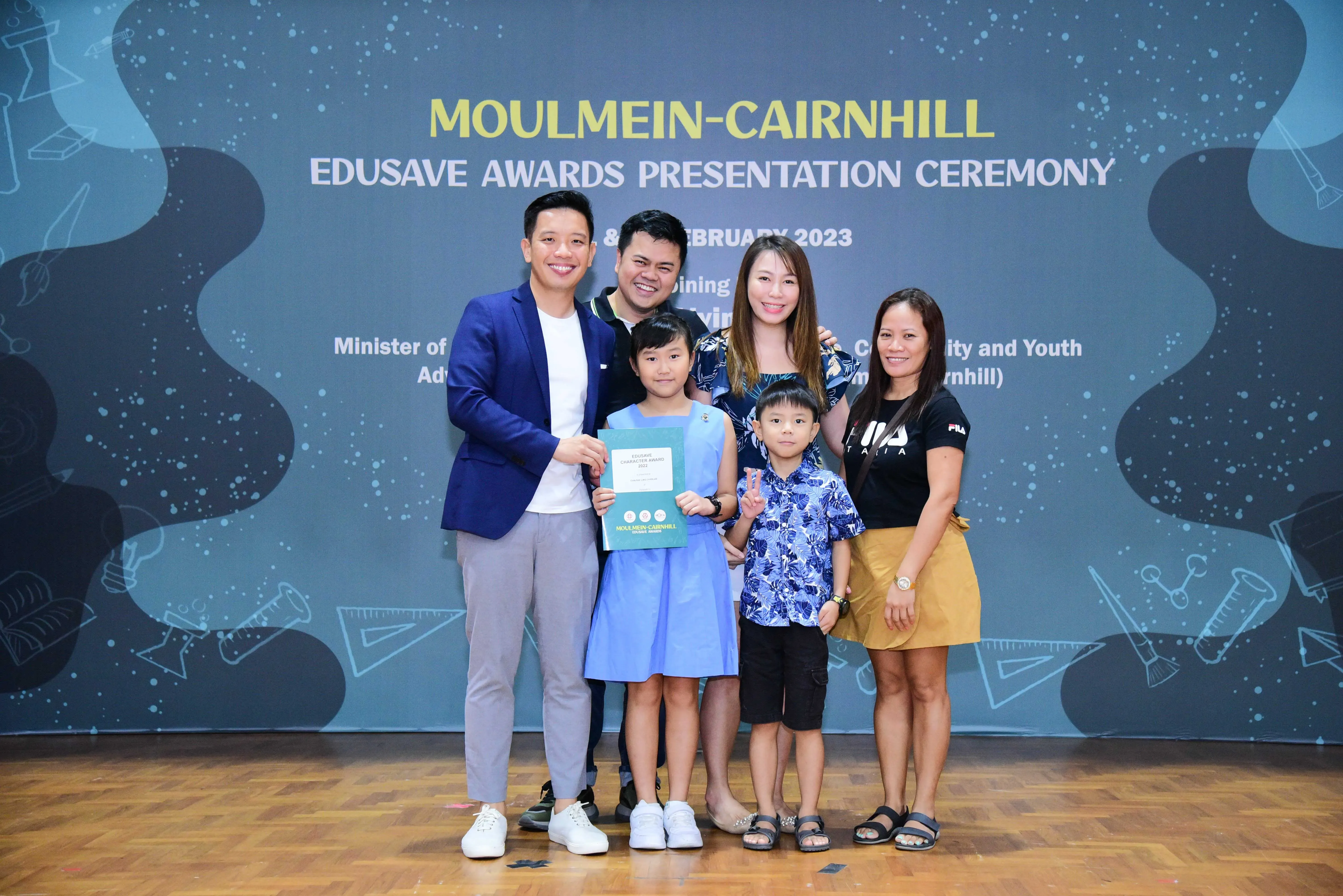 A girl holds a certificate with four adults and a boy at the Moulmein-Cairnhill Edusave Awards Ceremony, February 2023.