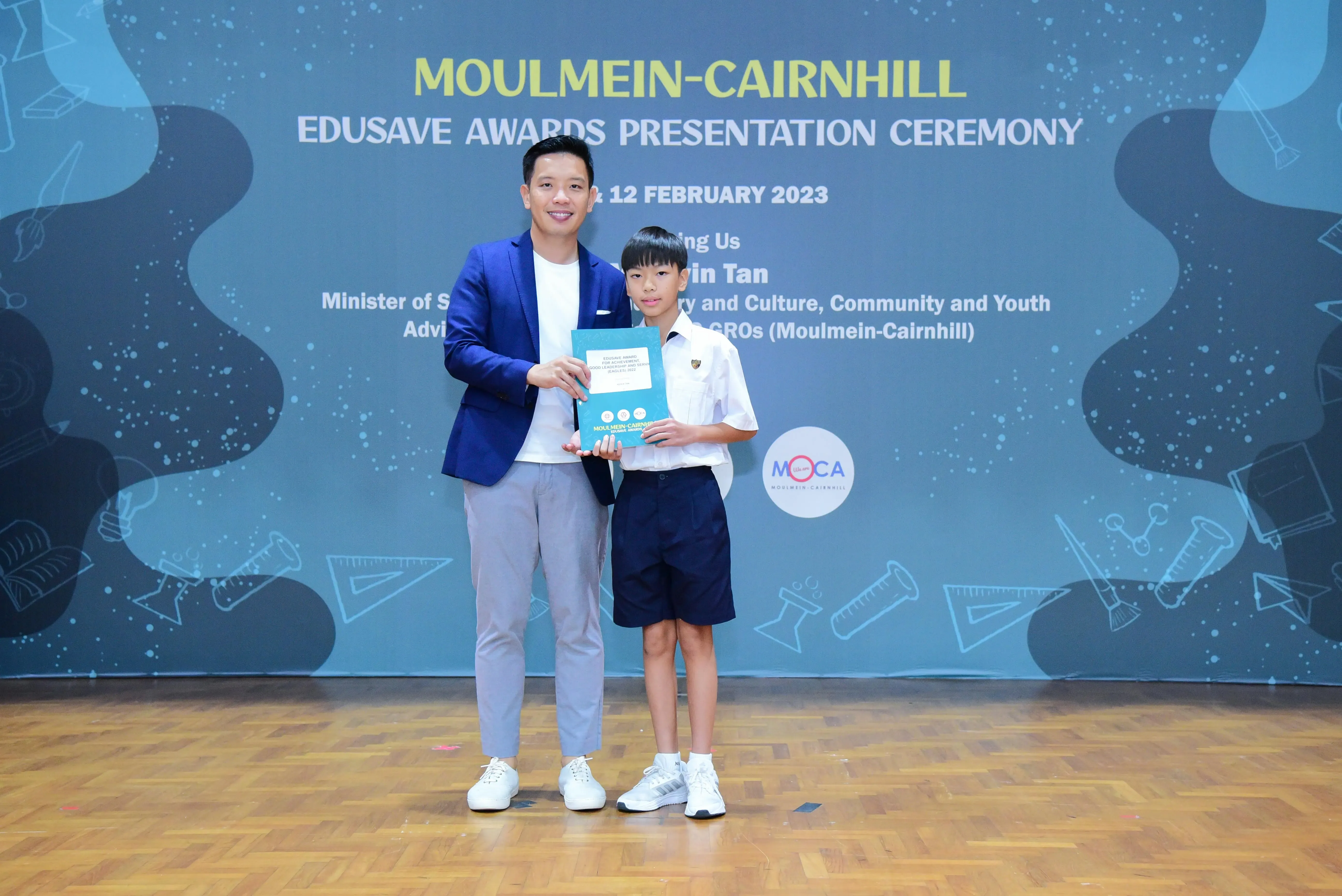 An adult in a blue blazer and a young student in uniform hold a certificate at the Moulmein-Cairnhill Edusave Awards 2023.