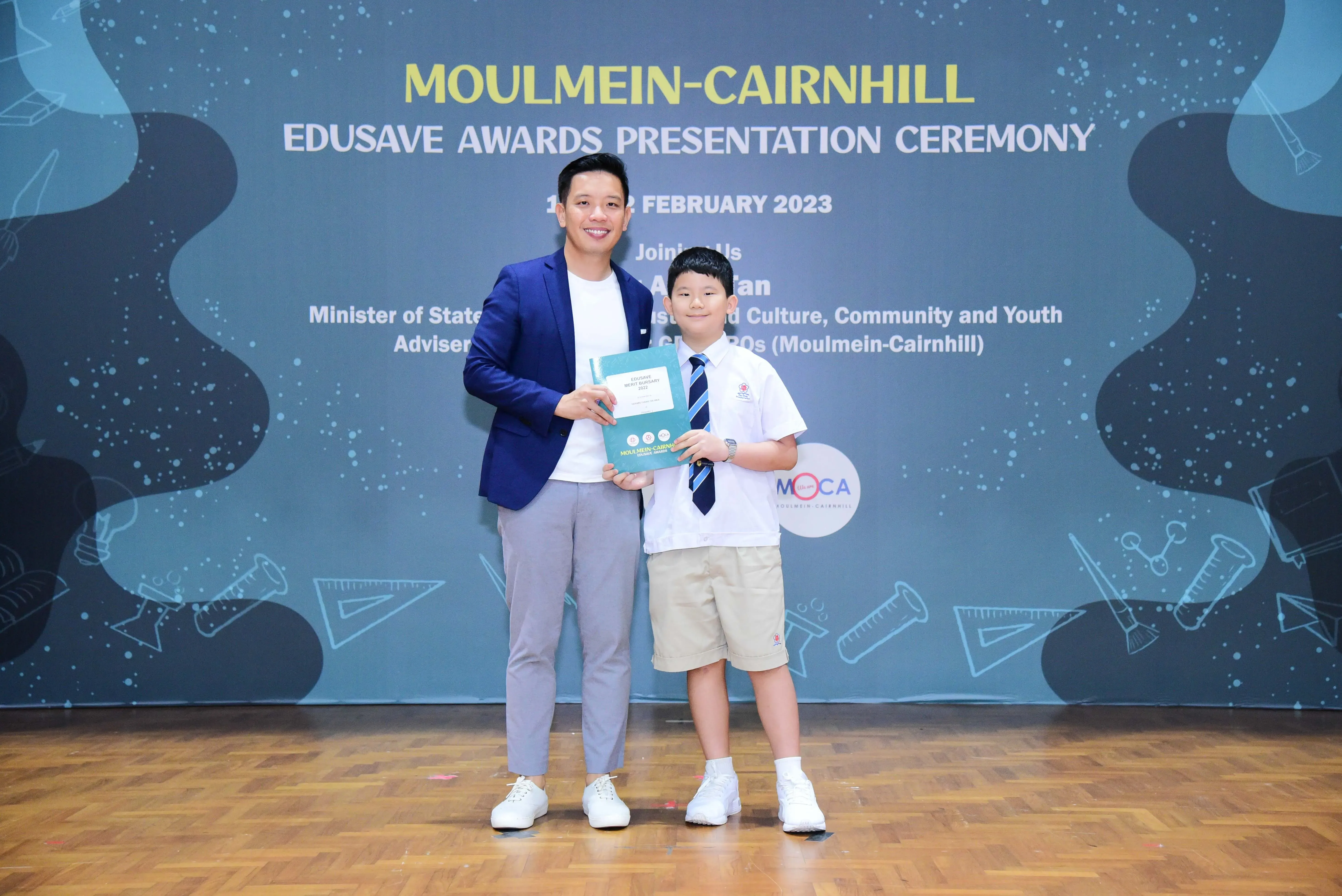 Adult in blue blazer and young boy in school uniform hold an award at Moulmein-Cairnhill Edusave Awards Ceremony 2023.