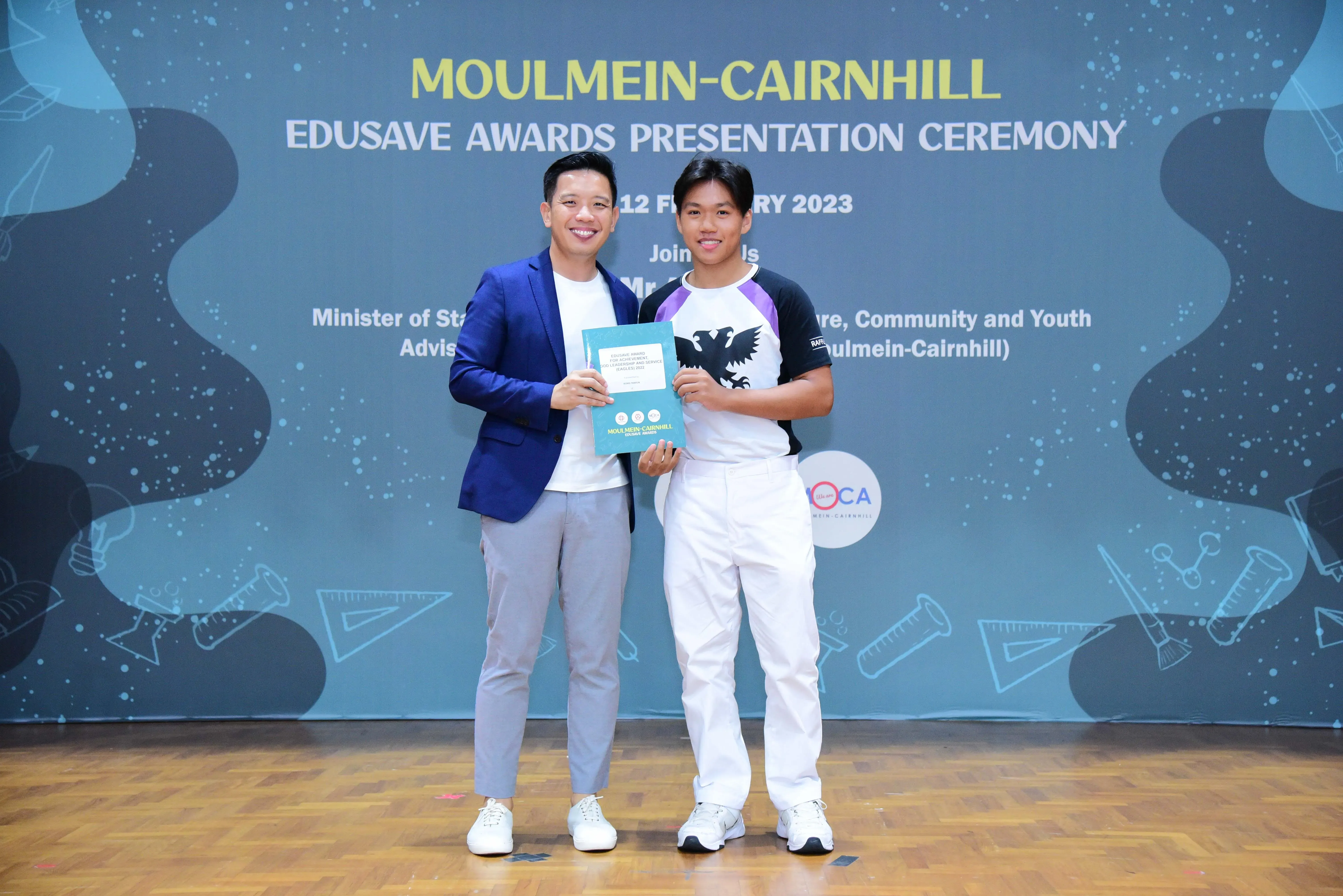 Two men smile holding an award booklet at the Moulmein-Cairnhill Edusave Awards Presentation Ceremony 2023.