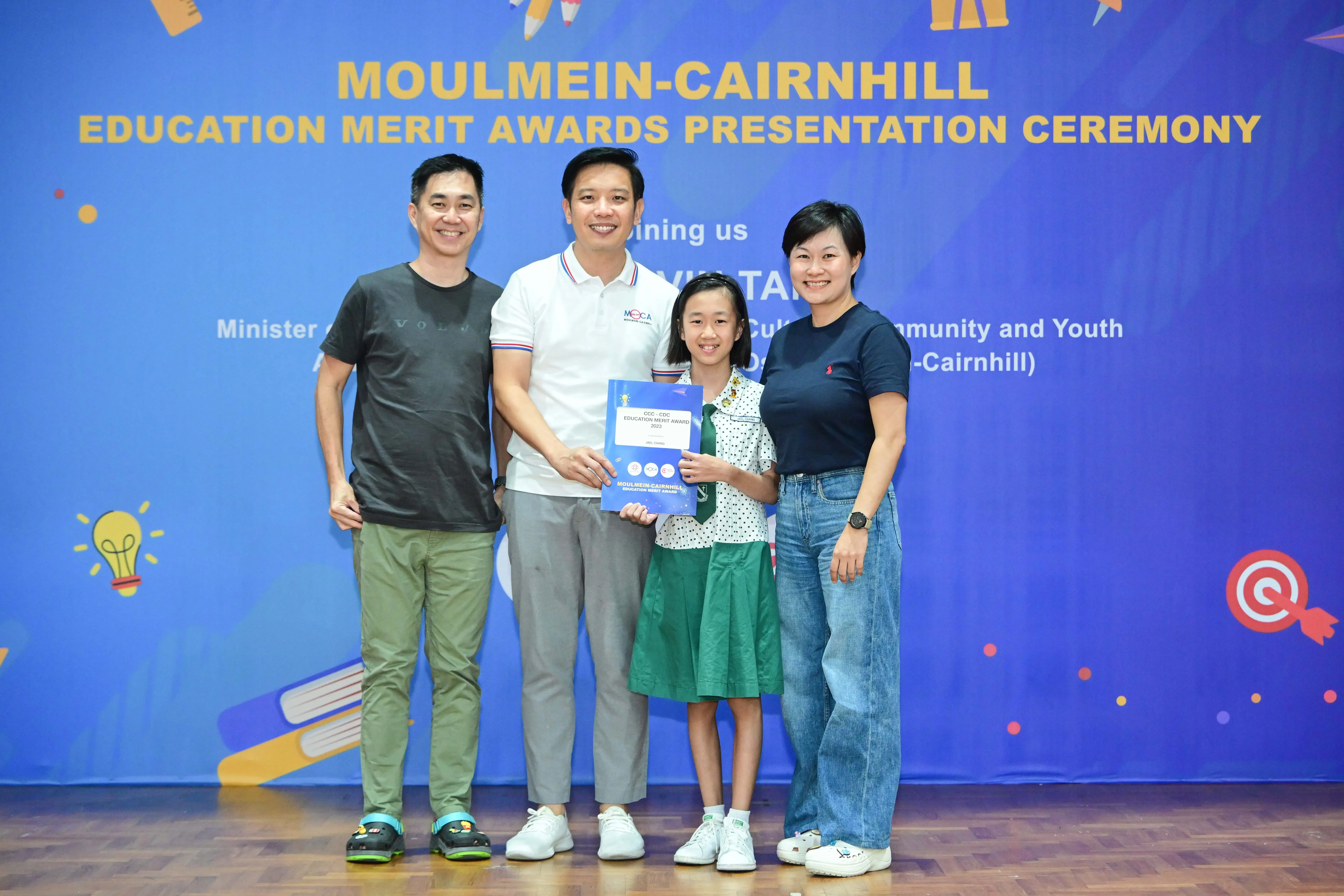 Four people pose before a Moulmein-Cairnhill Education Merit Awards banner, with a girl holding a blue certificate.