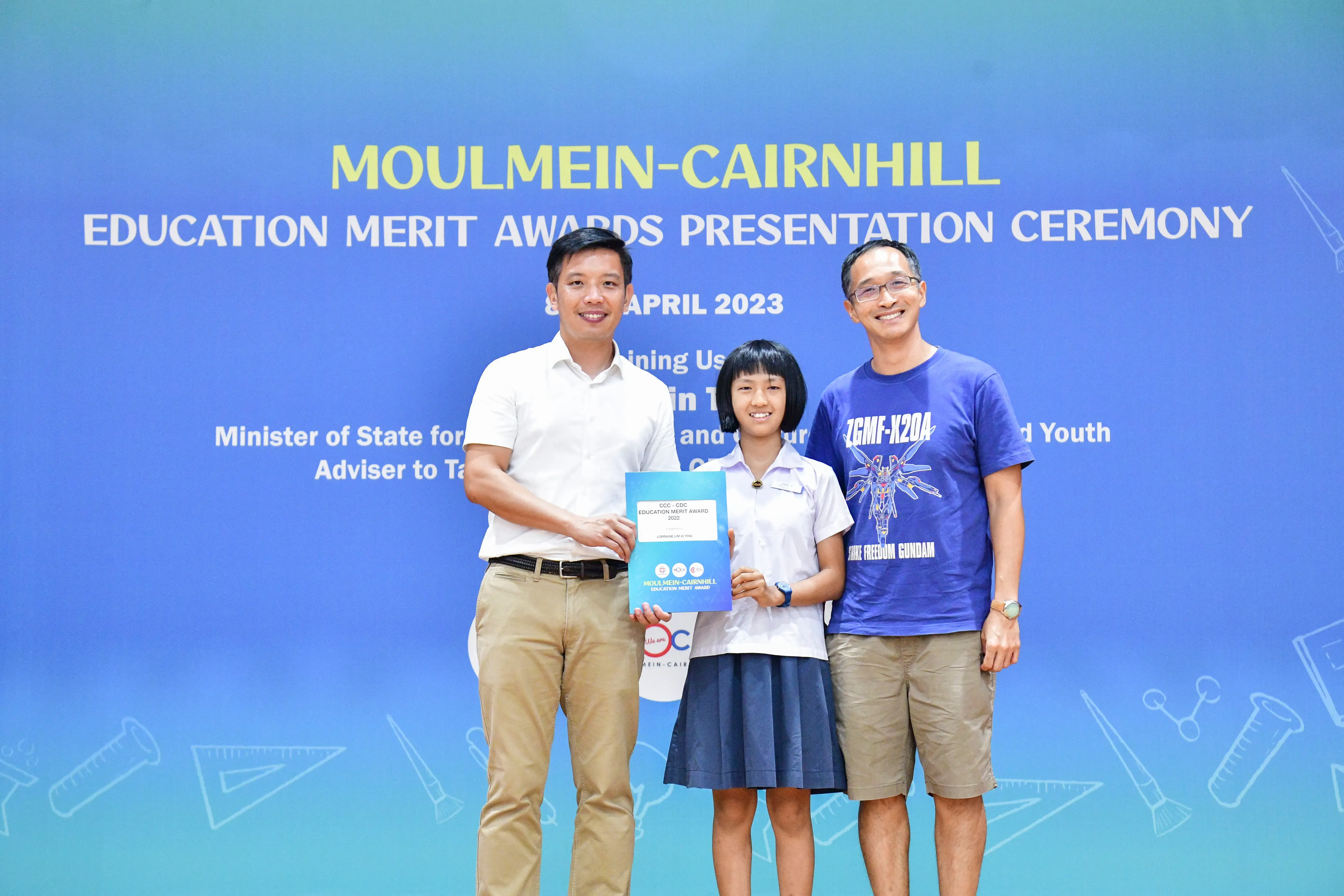 Three people pose before a Moulmein-Cairnhill Education Merit Awards Presentation Ceremony banner, girl holds certificate.