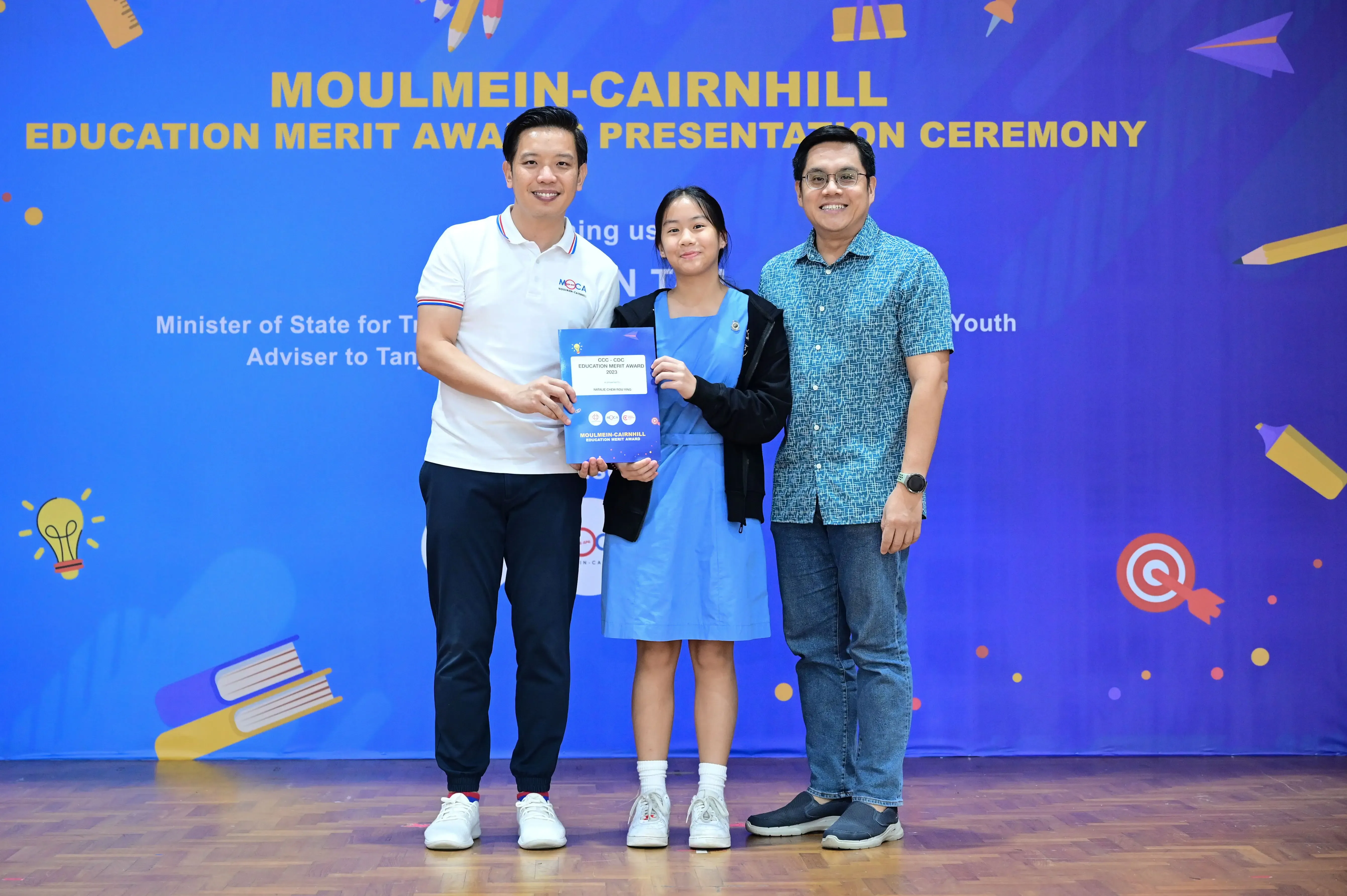 Three people pose at the Moulmein-Cairnhill Education Merit Award Presentation Ceremony, with a girl holding a certificate.