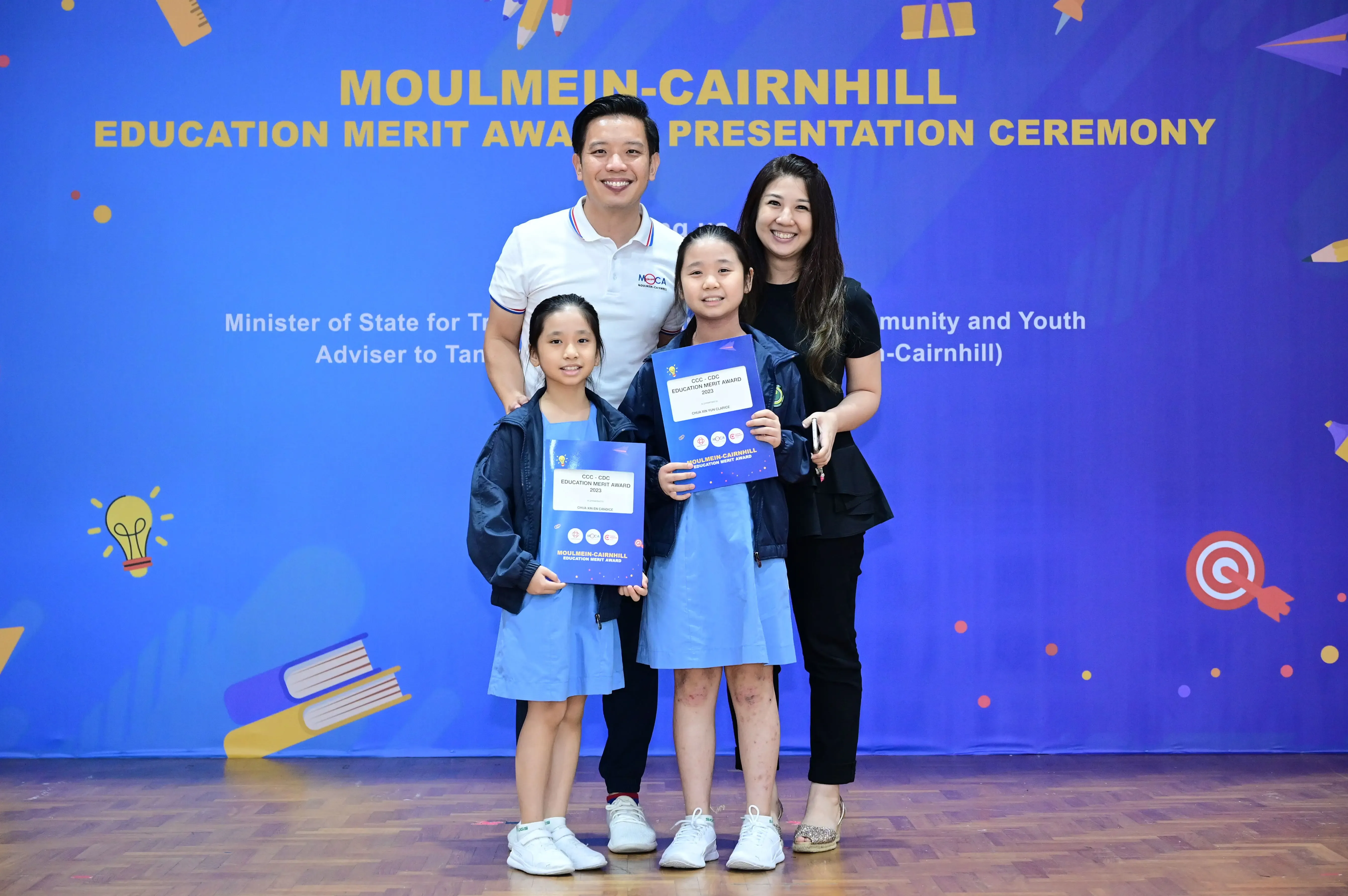 Two adults and two girls holding award certificates pose before a Moulmein-Cairnhill Education Merit Award banner.