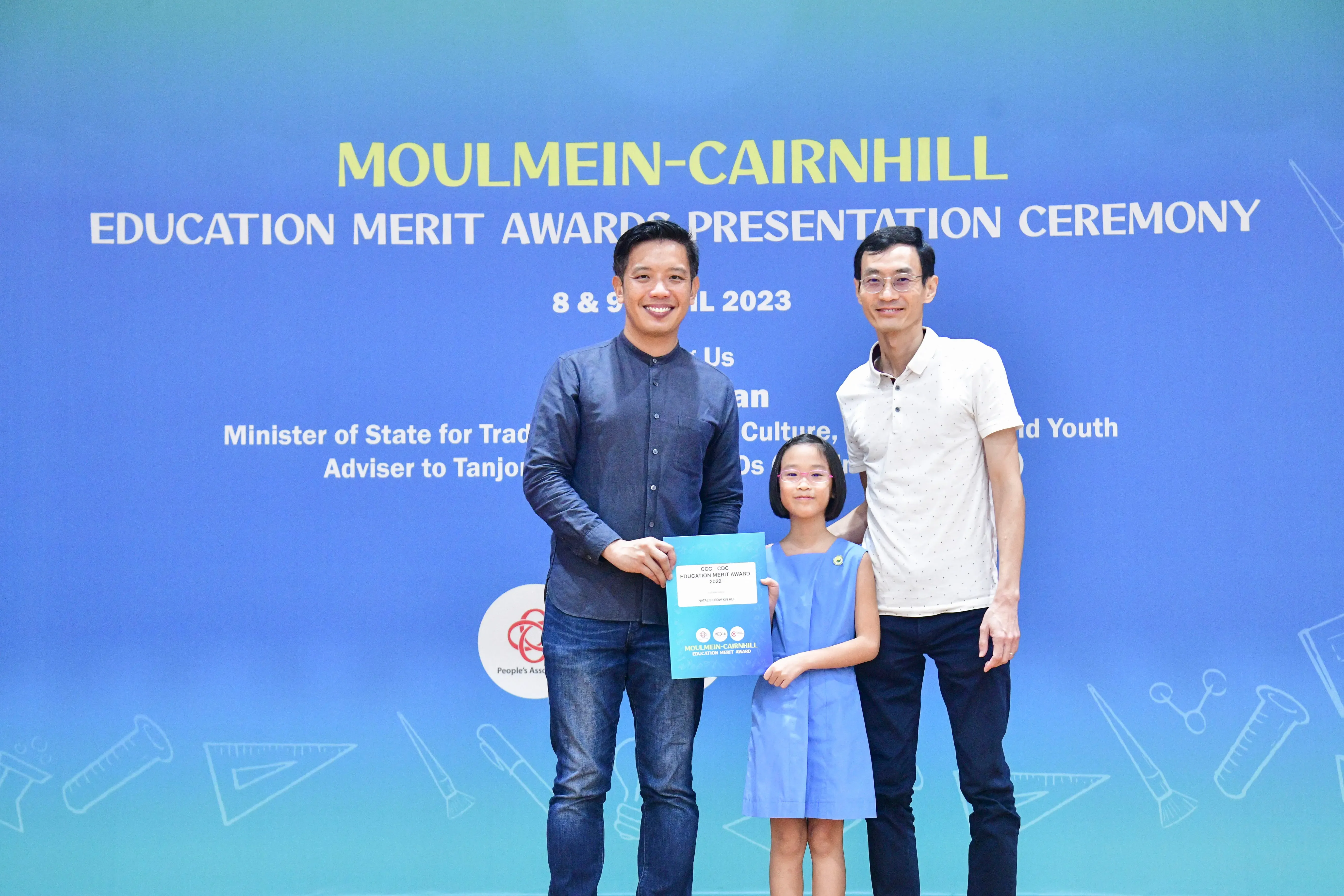 Two men and a young girl holding a certificate pose before a Moulmein-Cairnhill Education Merit Awards banner.