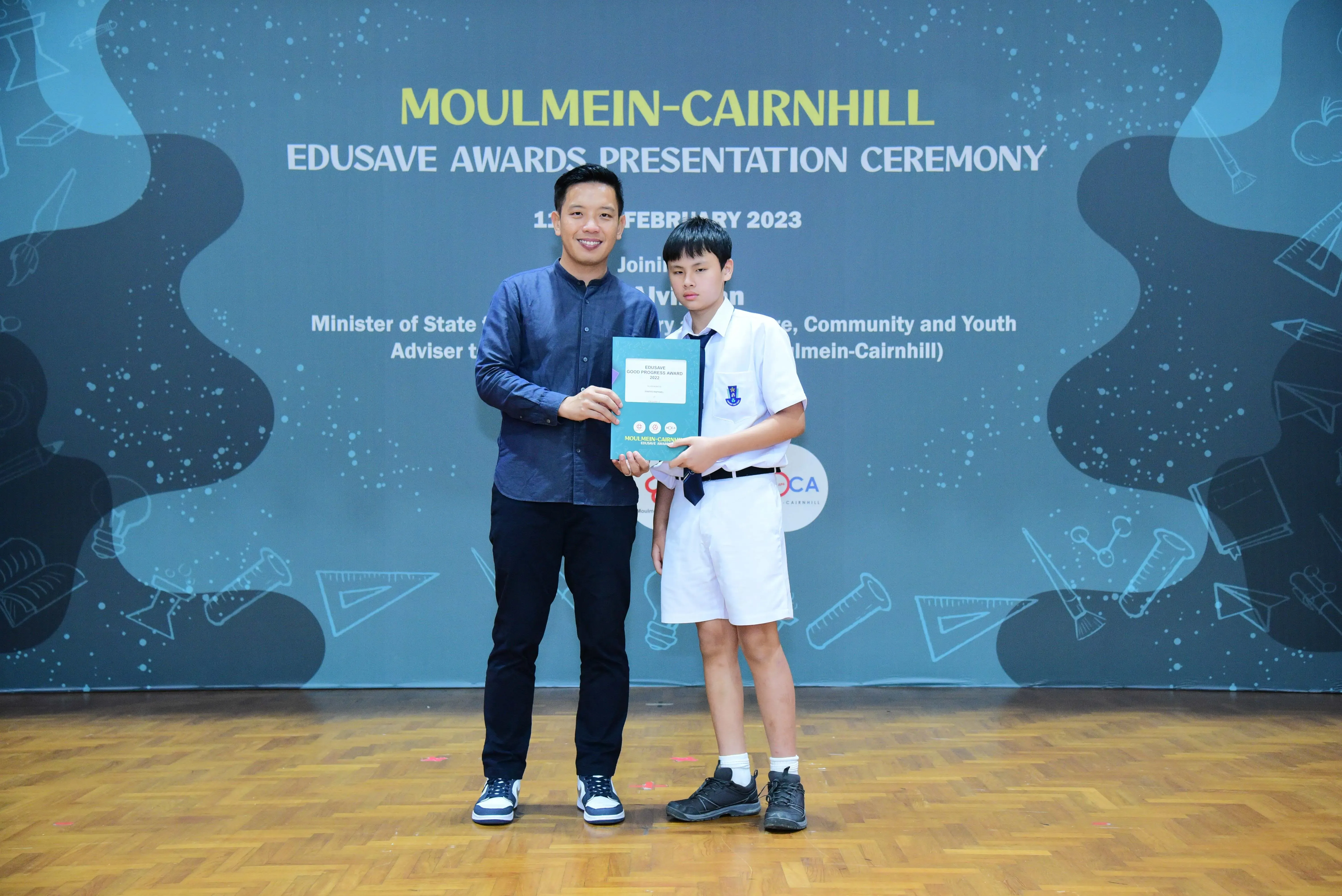An adult and a student in uniform hold a certificate at the Moulmein-Cairnhill Edusave Awards Ceremony, February 2023.