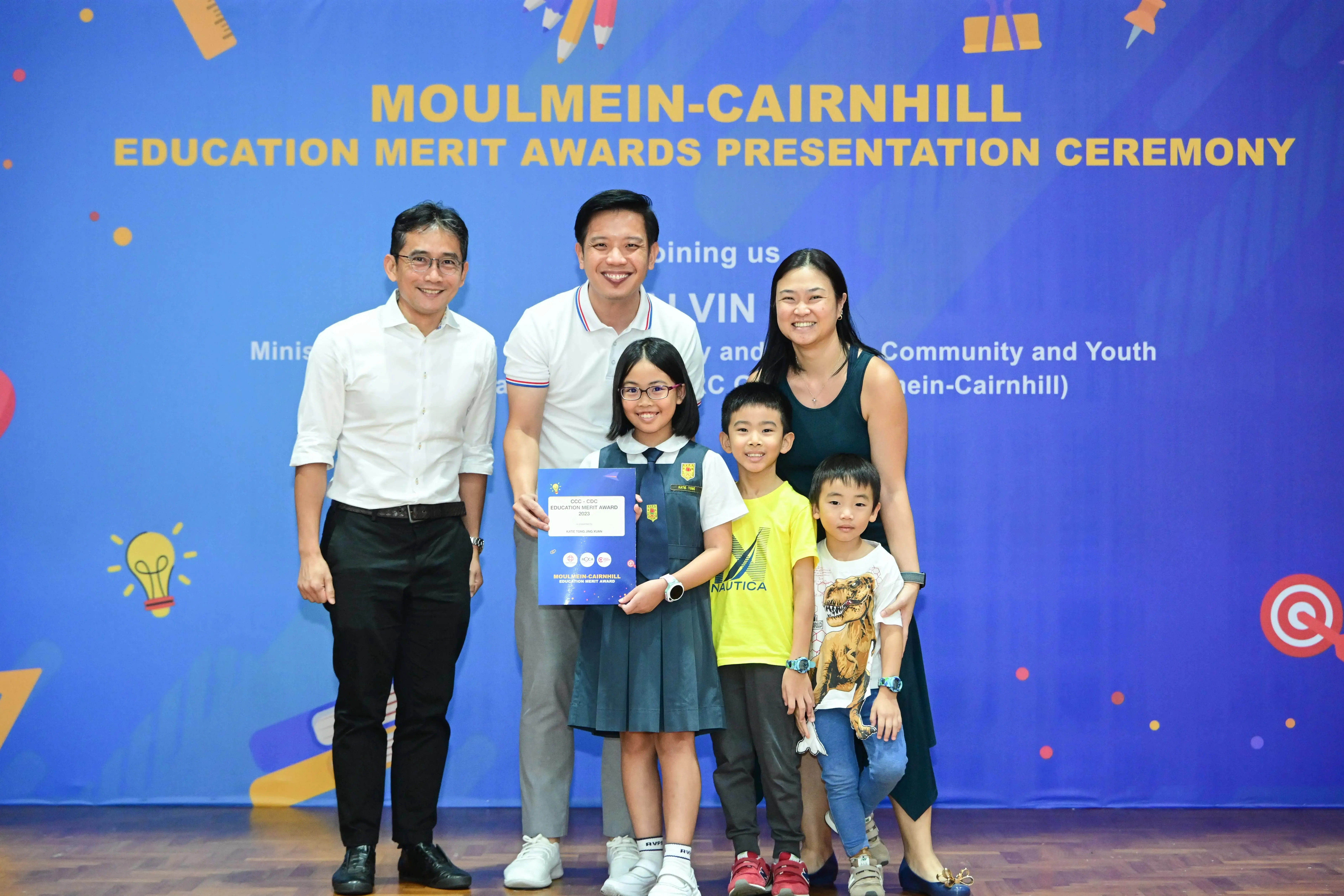 A girl in school uniform holds an award certificate, posing with two men, a woman, and two boys at an education merit ceremony.
