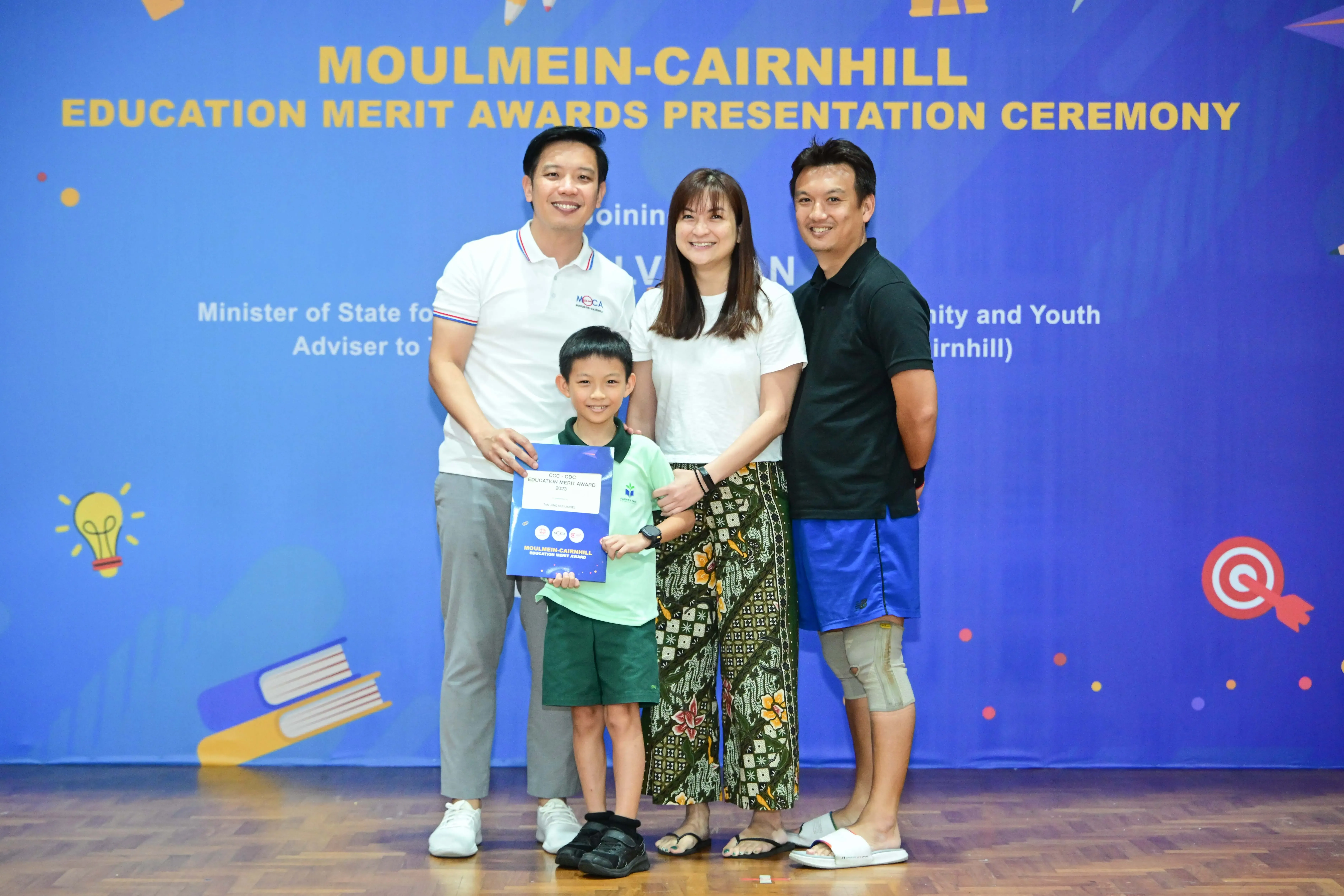 Three adults and a young boy holding a certificate pose before a Moulmein-Cairnhill Education Merit Awards banner.