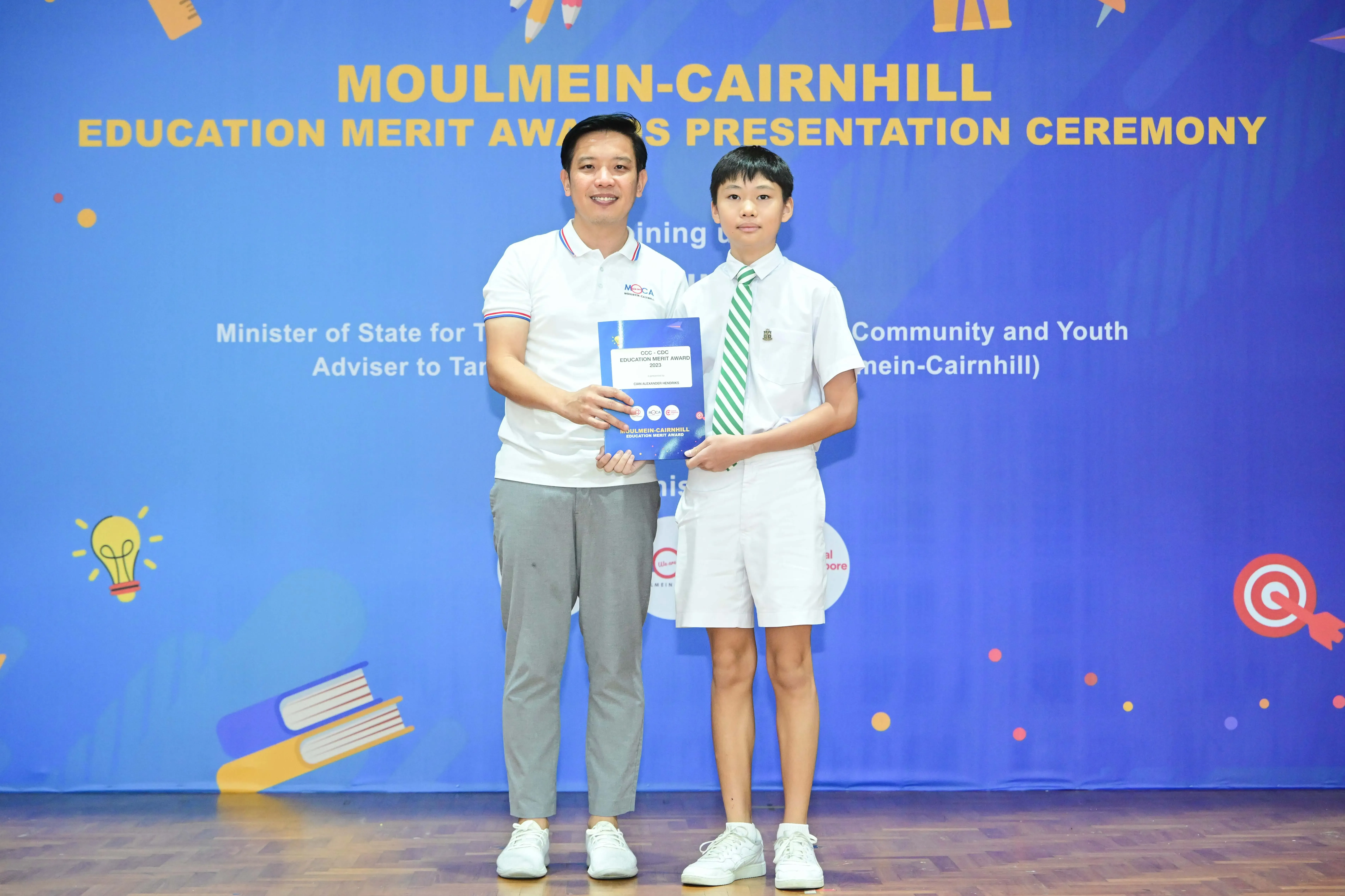 An adult and a young student in uniform hold an award together at the Moulmein-Cairnhill Education Merit Awards ceremony.