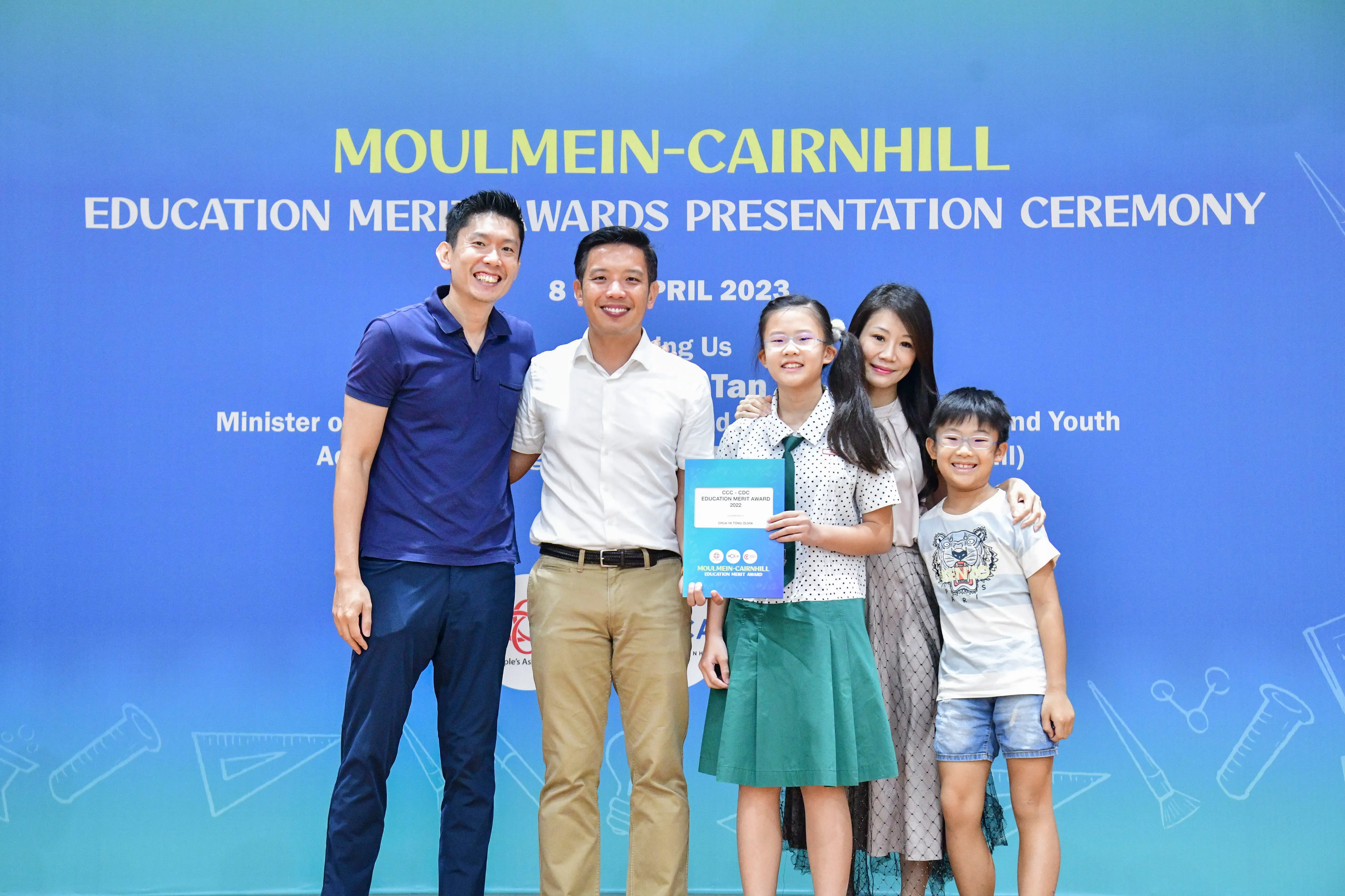Five people pose smiling before a blue Moulmein-Cairnhill Education Merit Awards Presentation Ceremony banner dated April 8, 2023.
