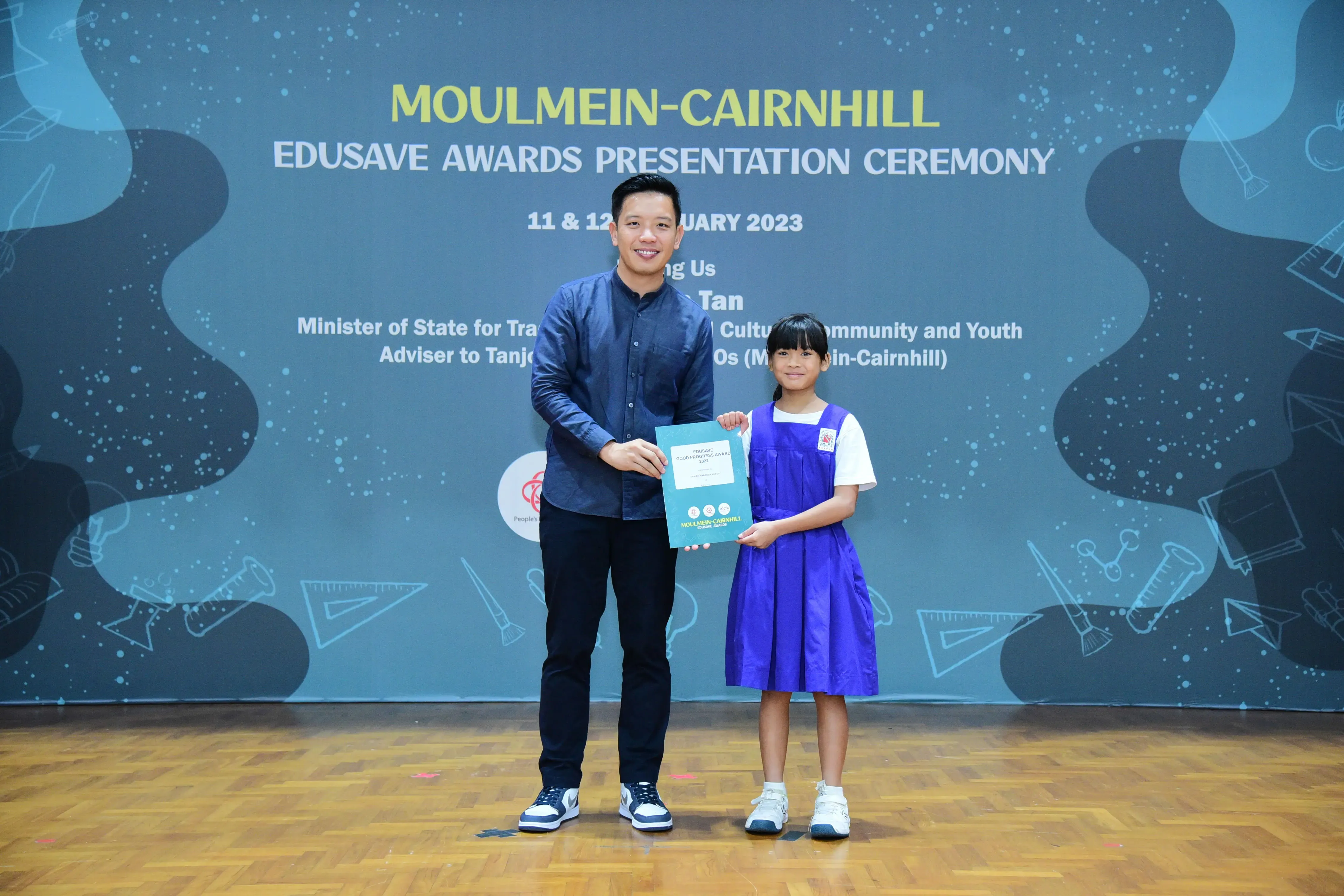 A man and a young girl in school uniform hold a certificate at the Moulmein-Cairnhill Edusave Awards Ceremony 2023.