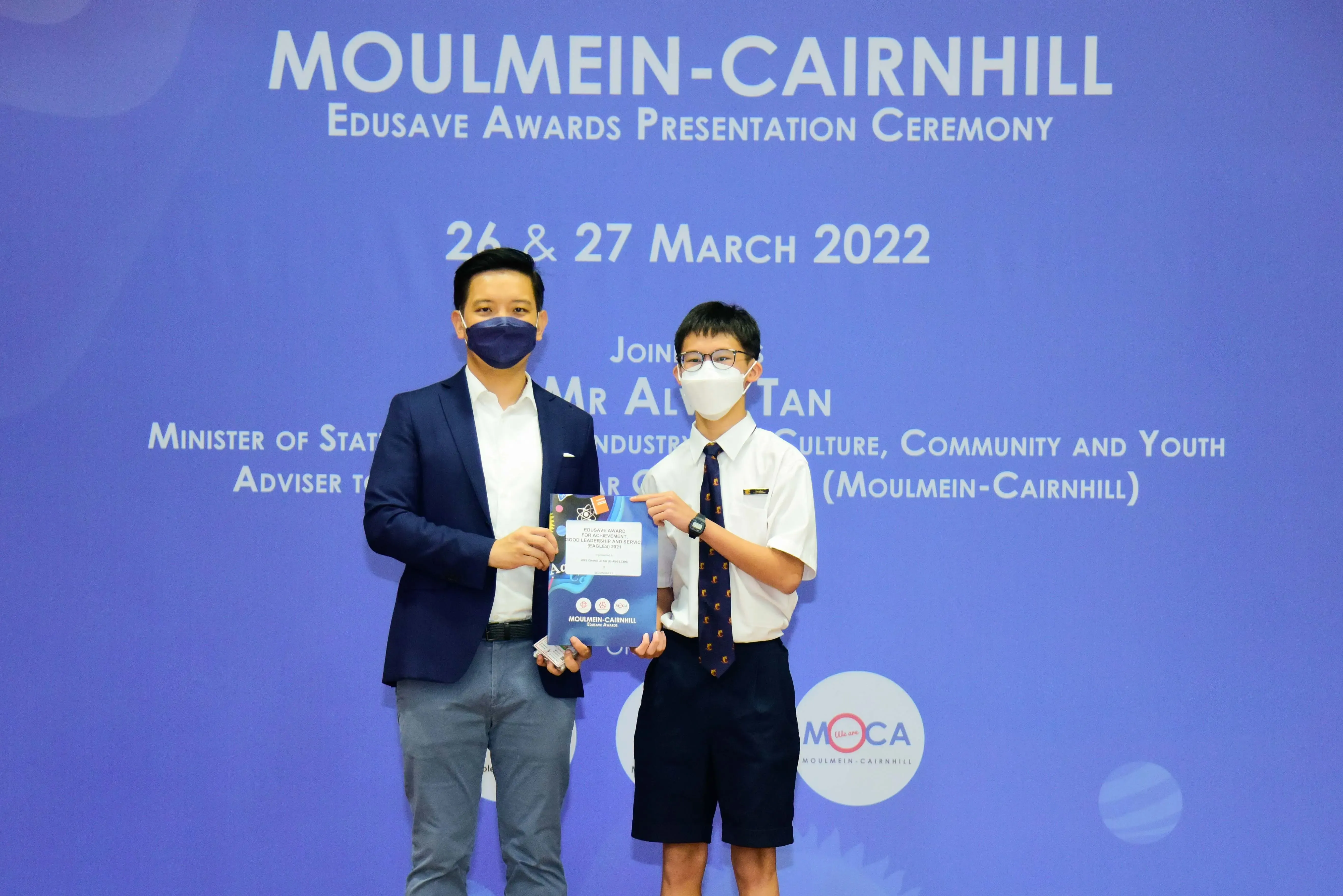 Two masked individuals hold an award at the Moulmein-Cairnhill Edusave Awards Presentation Ceremony, March 2022.