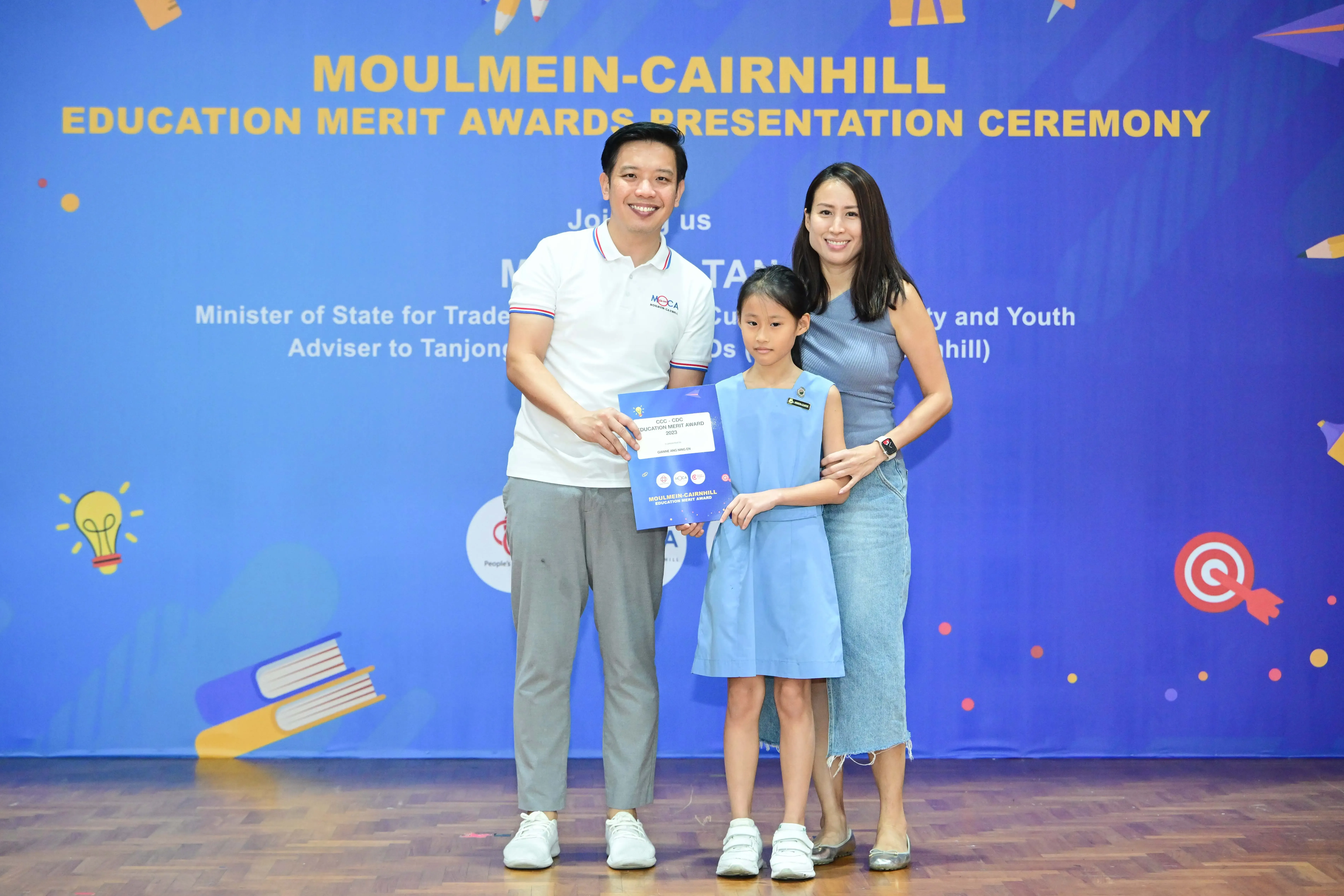 A man, young girl holding an award certificate, and woman pose at the Moulmein-Cairnhill Education Merit Awards ceremony.