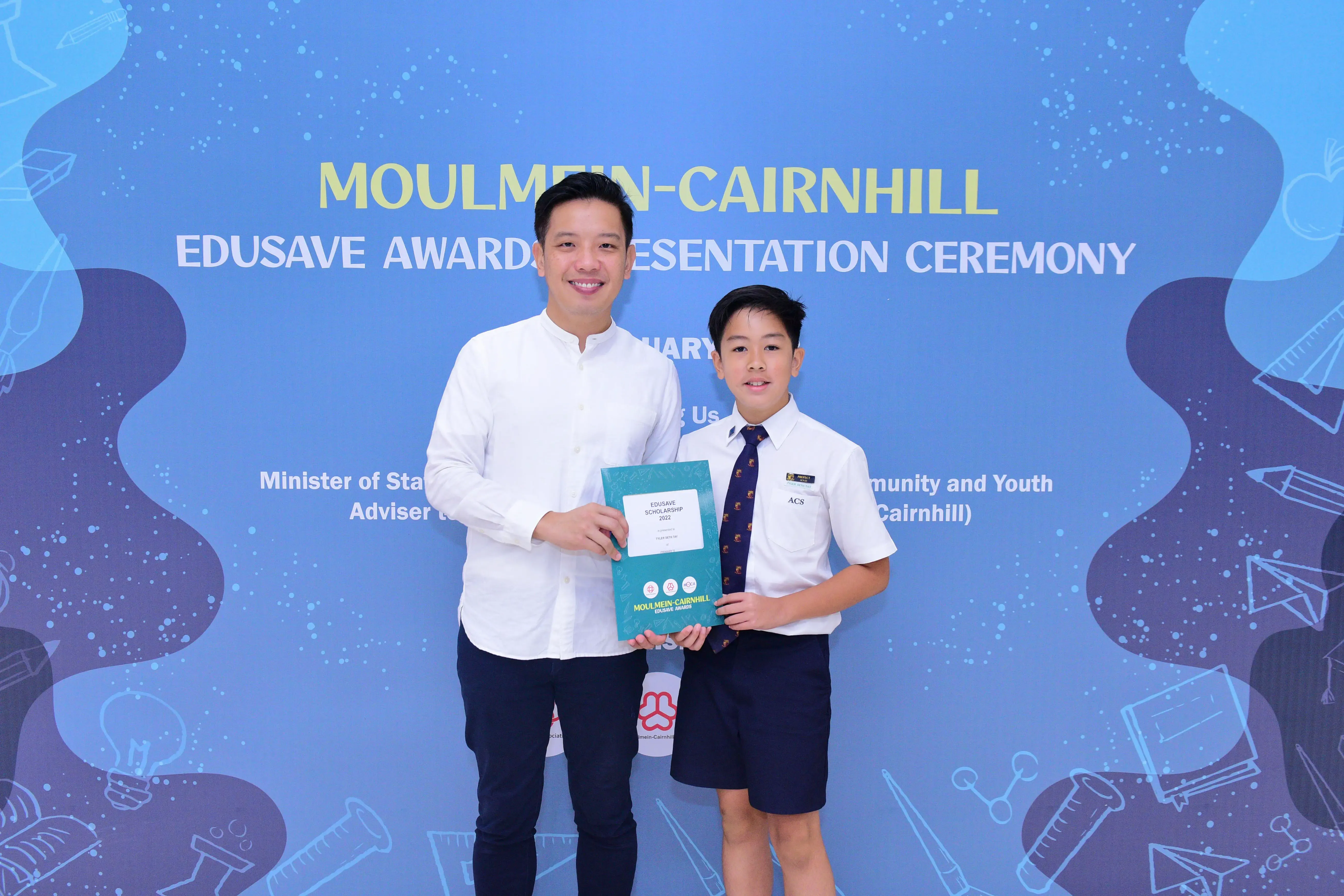 A man and a young boy in school uniform hold an Edusave Scholarship certificate at the Moulmein-Cairnhill Awards Ceremony.