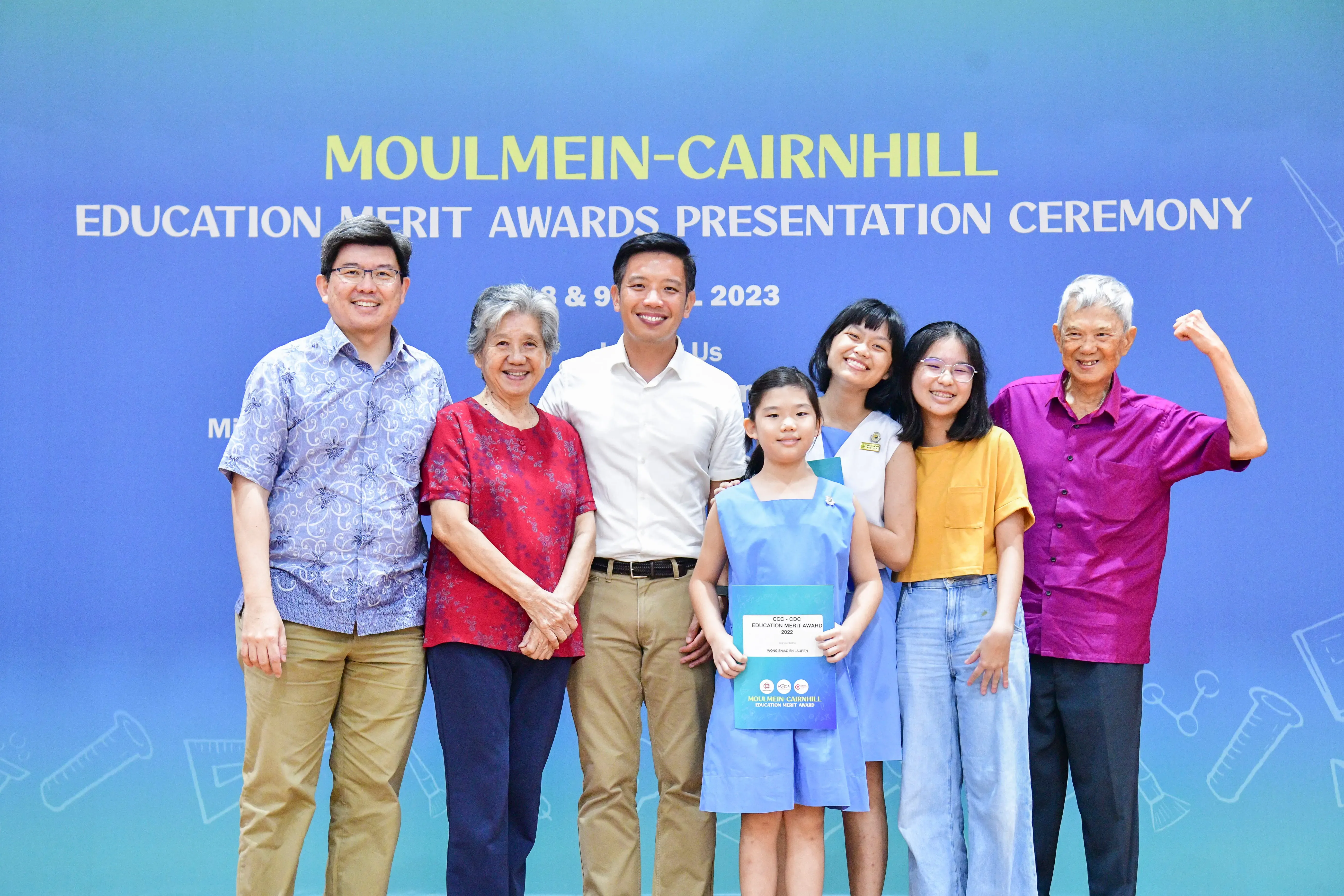 Seven people pose smiling at the 2023 Moulmein-Cairnhill Education Merit Awards Presentation Ceremony backdrop.