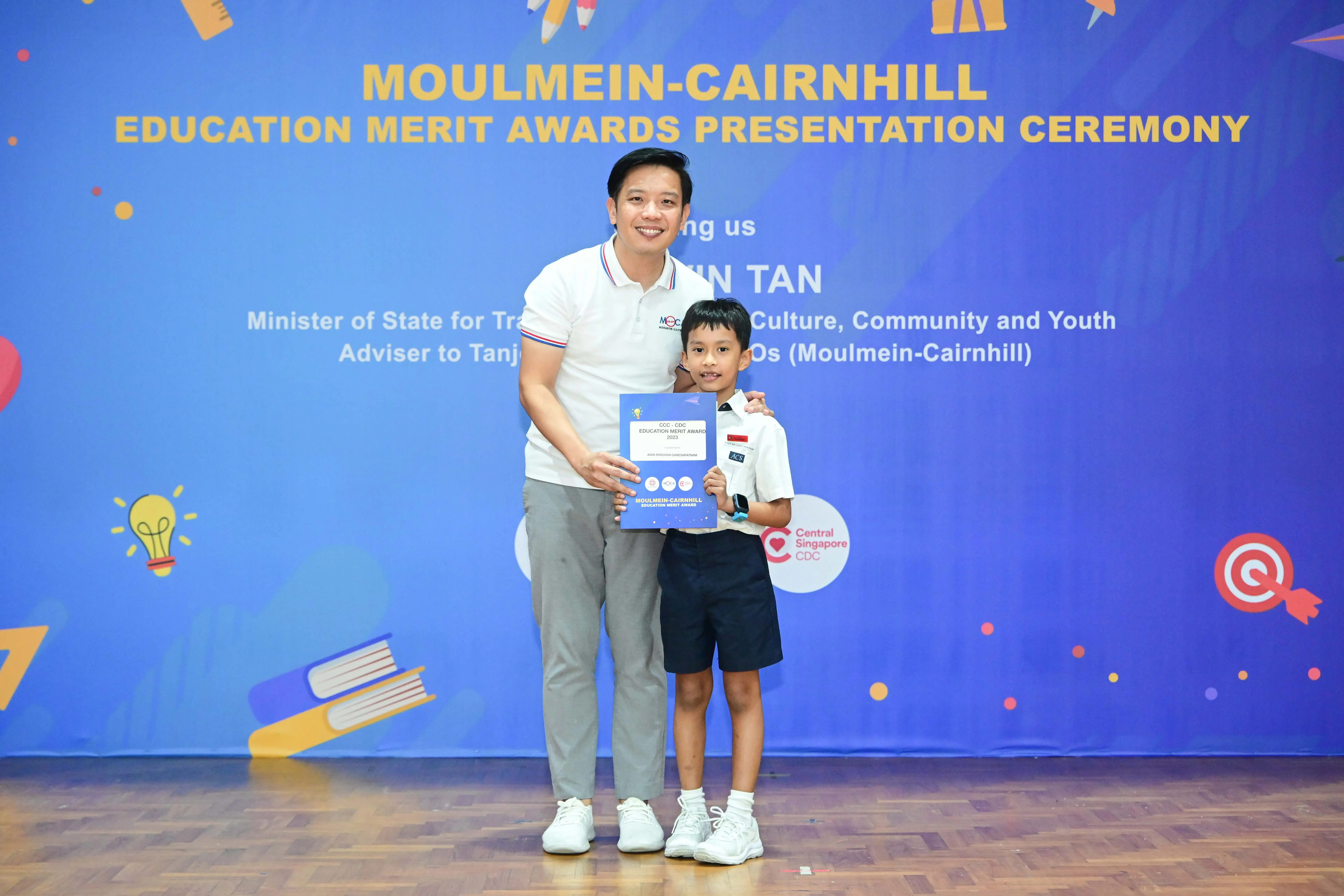 A man and a young boy hold a certificate in front of a Moulmein-Cairnhill Education Merit Awards banner.