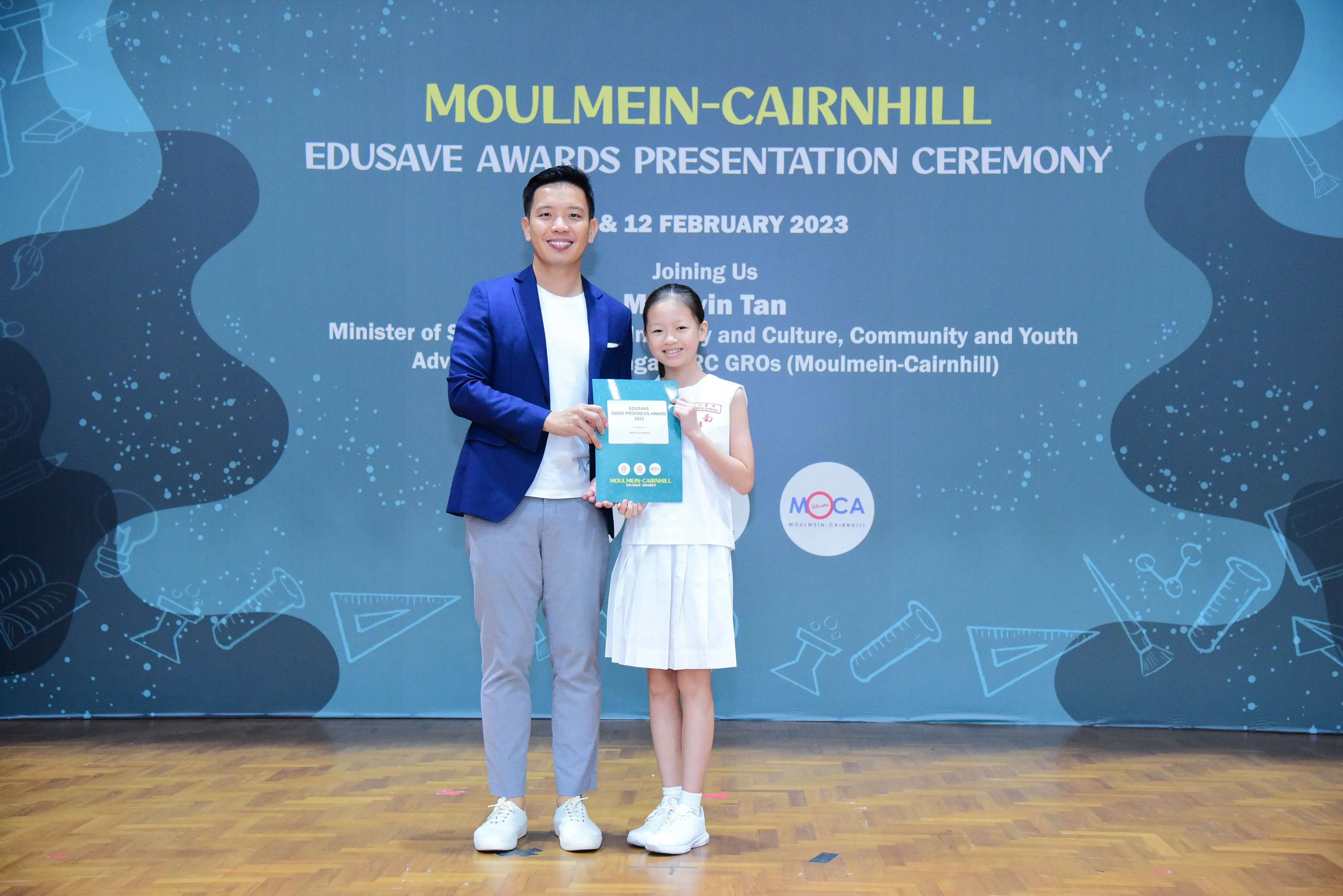 Man in blue blazer and young girl in white dress hold a certificate at Moulmein-Cairnhill Edusave Awards Ceremony 2023.
