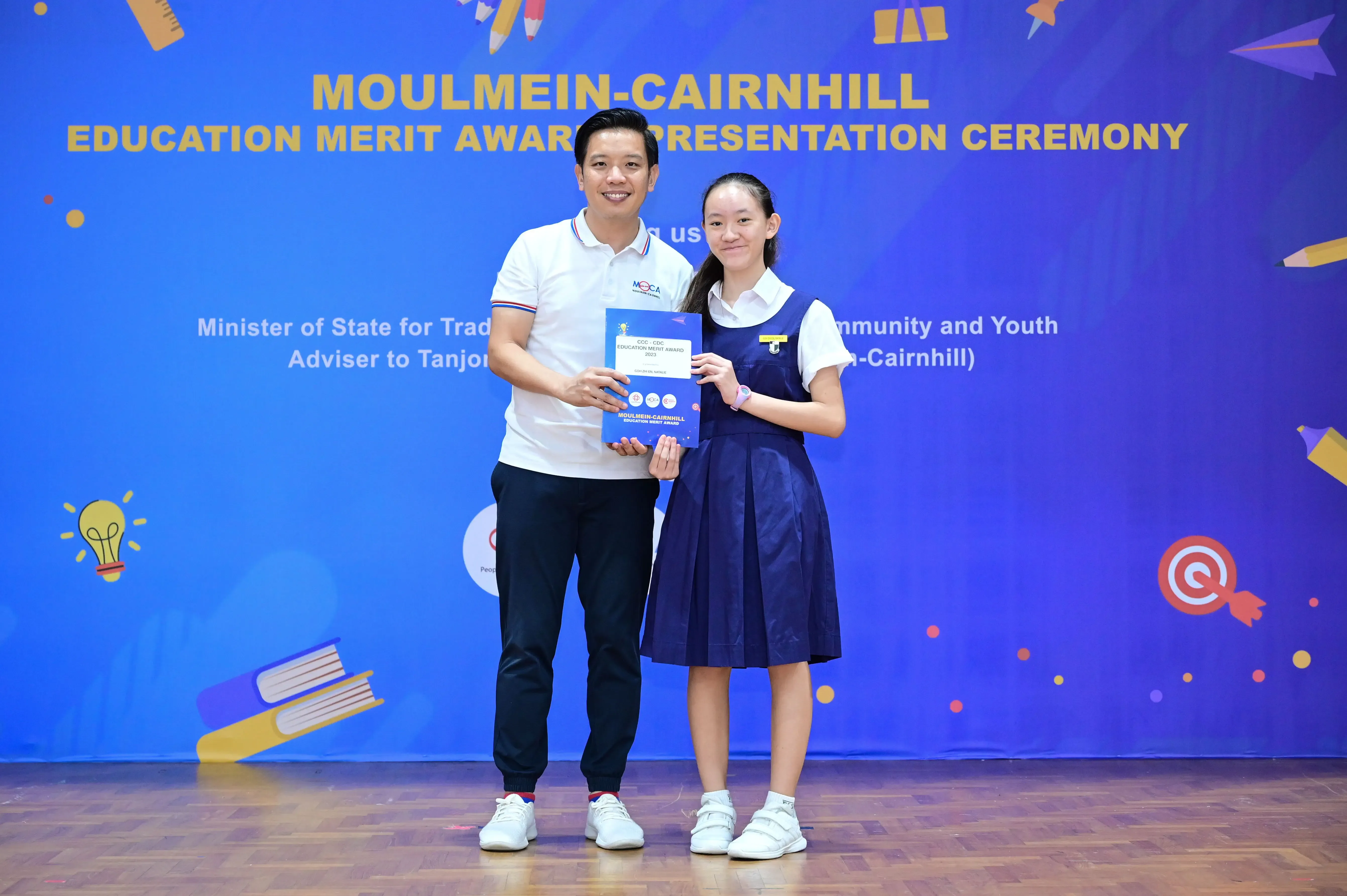 A man and a young girl in school uniform hold an award certificate at the Moulmein-Cairnhill Education Merit Award ceremony.