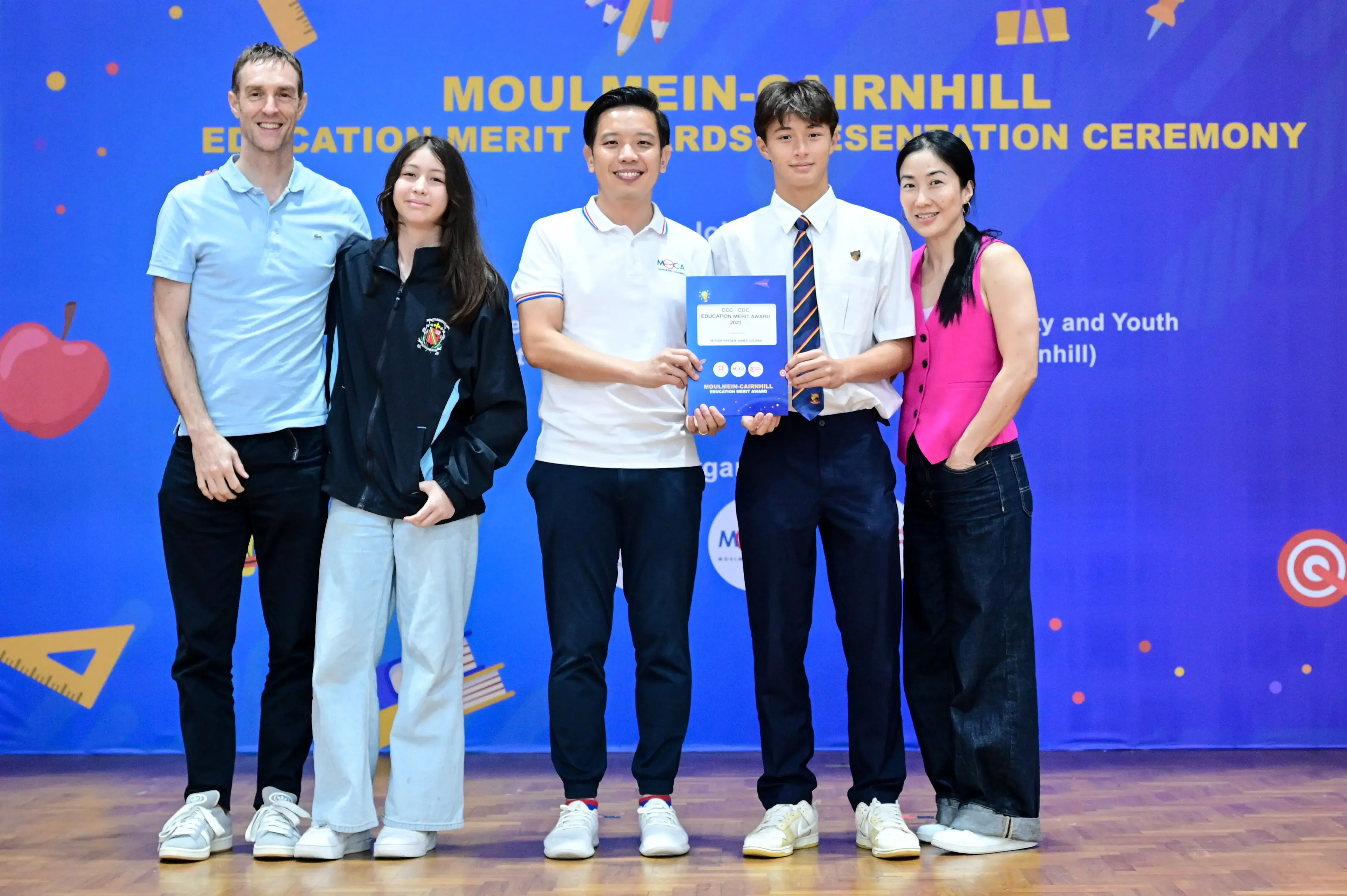 Five people pose on stage at the Moulmein-Cairnhill Education Merit Awards Presentation Ceremony holding a certificate.
