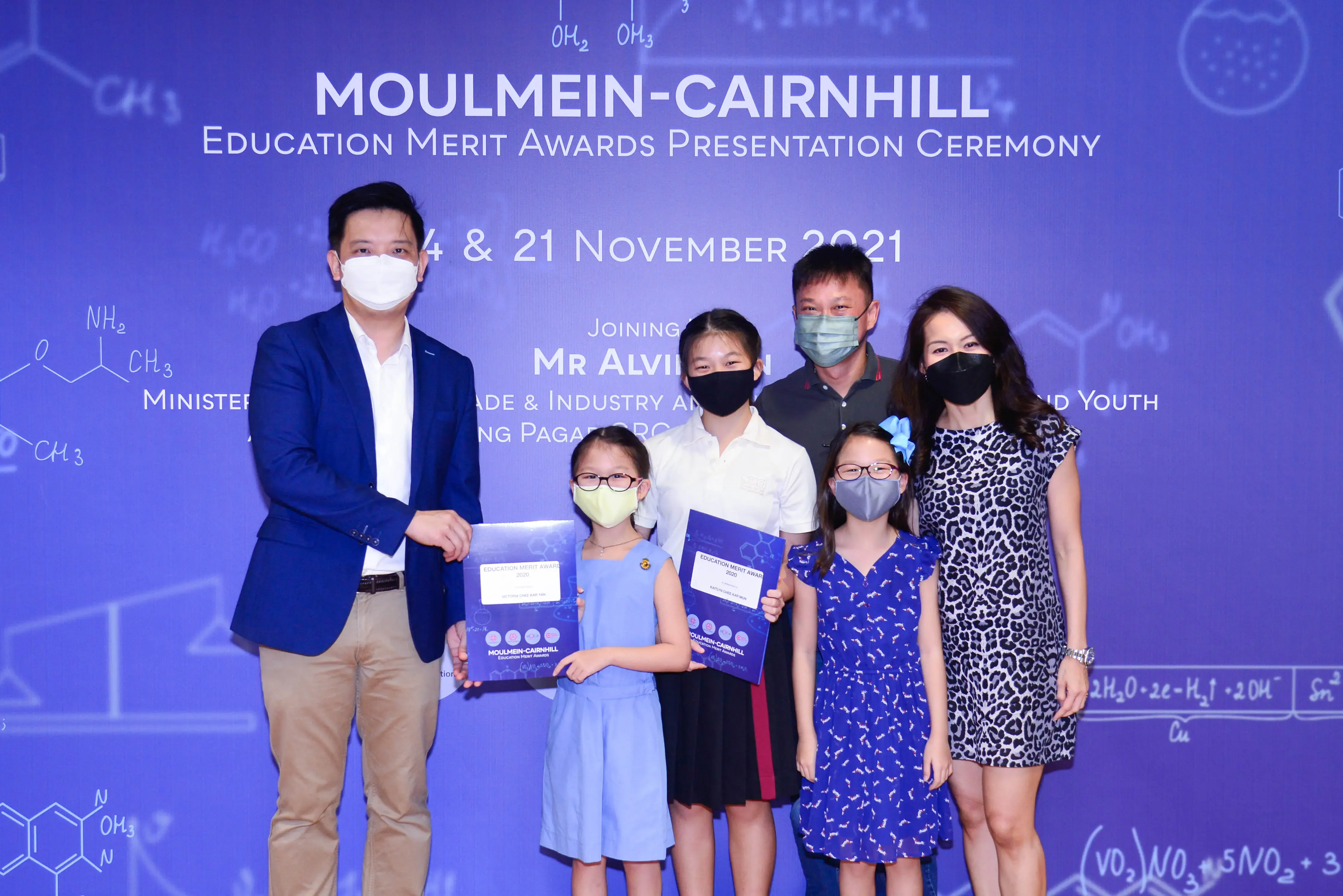 Six masked people pose at the Moulmein-Cairnhill Education Merit Awards Ceremony, November 2021, with children holding award certificates.