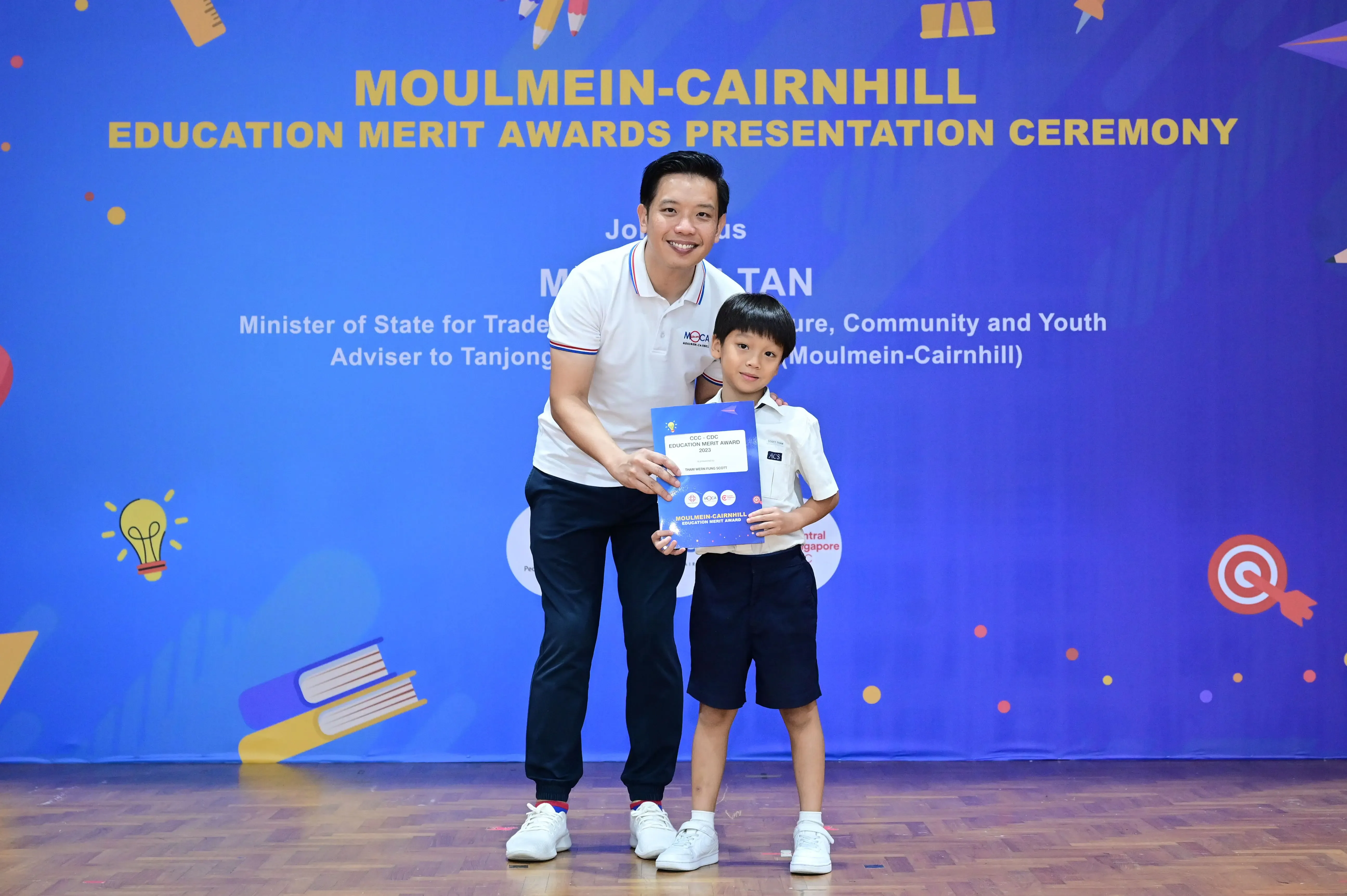 A man and a young boy hold a blue certificate at the Moulmein-Cairnhill Education Merit Awards Presentation Ceremony.