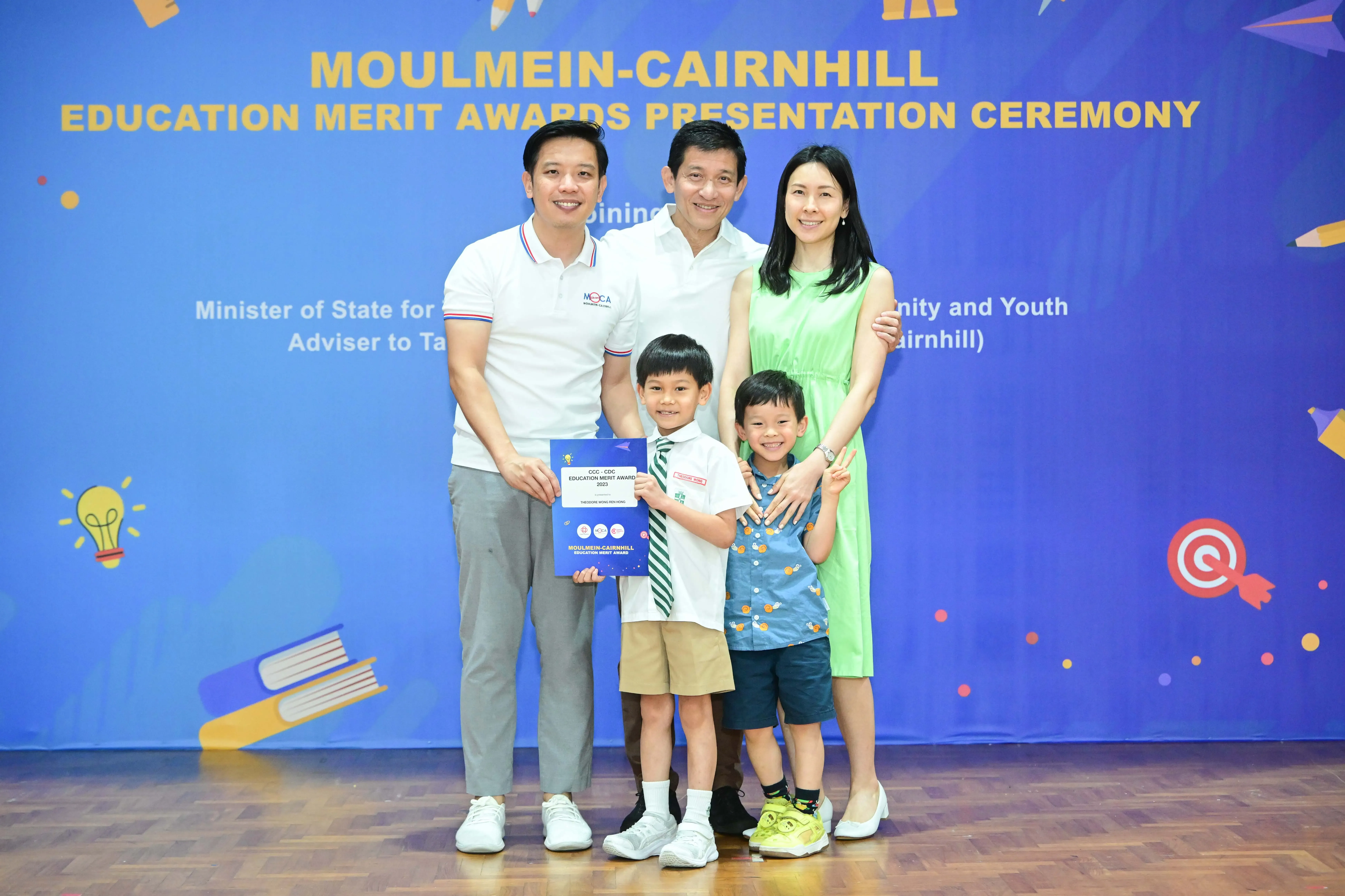 Five people pose before a Moulmein-Cairnhill Education Merit Awards banner, with a boy holding a blue certificate.