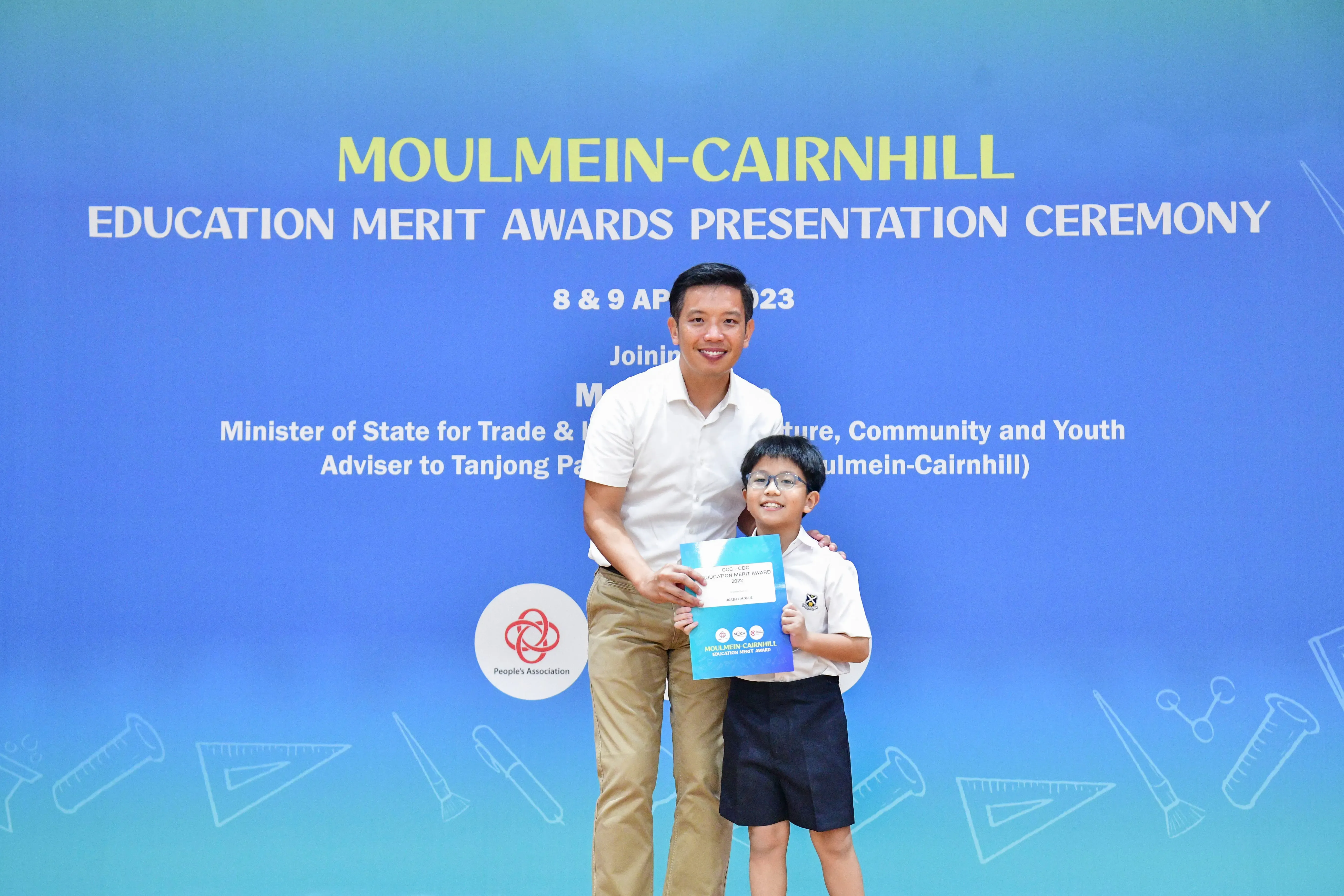 A man and a young boy holding an award certificate pose before a Moulmein-Cairnhill Education Merit Awards backdrop.
