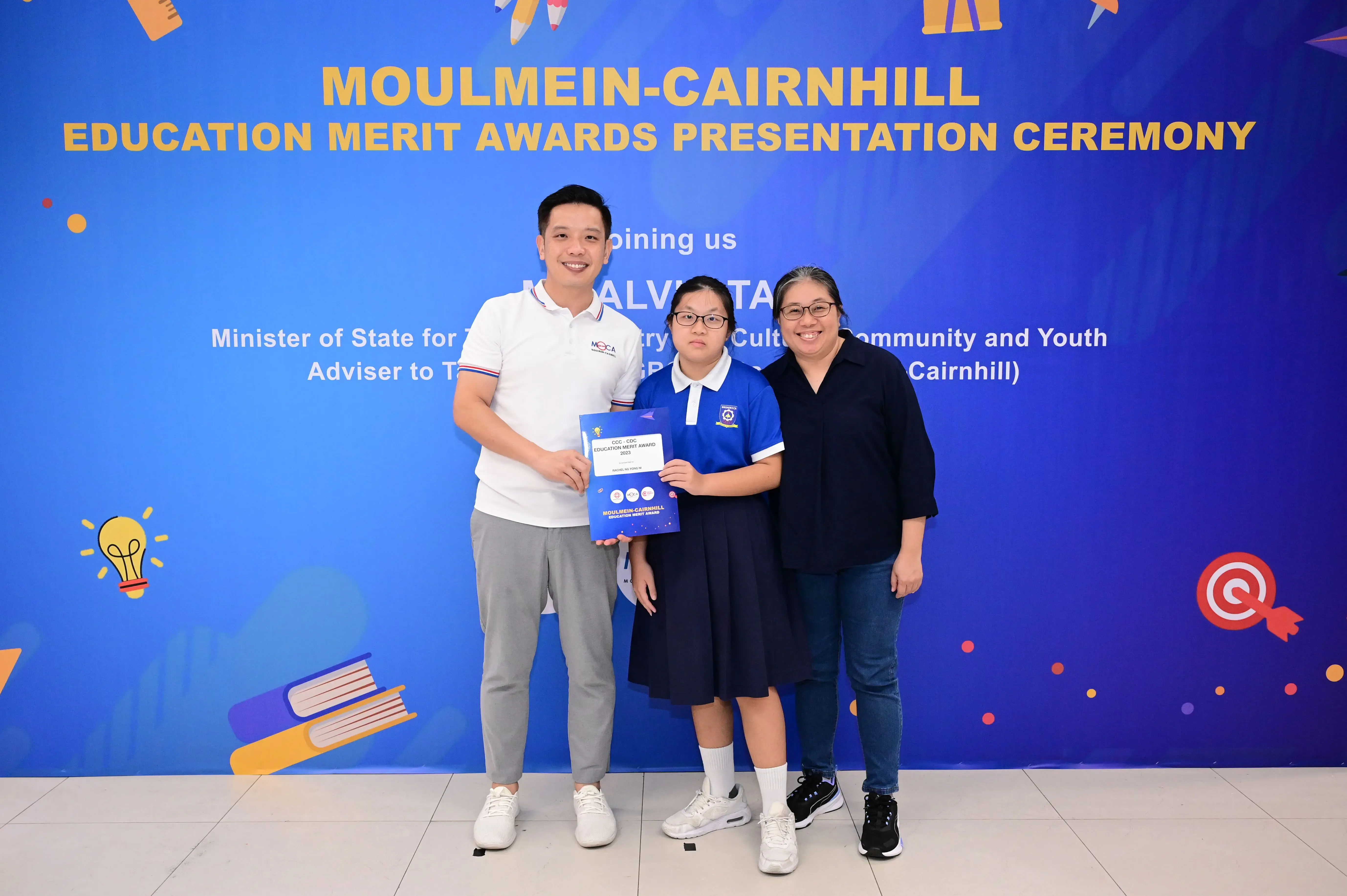 Three people pose before a Moulmein-Cairnhill Education Merit Awards banner, with a student holding a certificate.