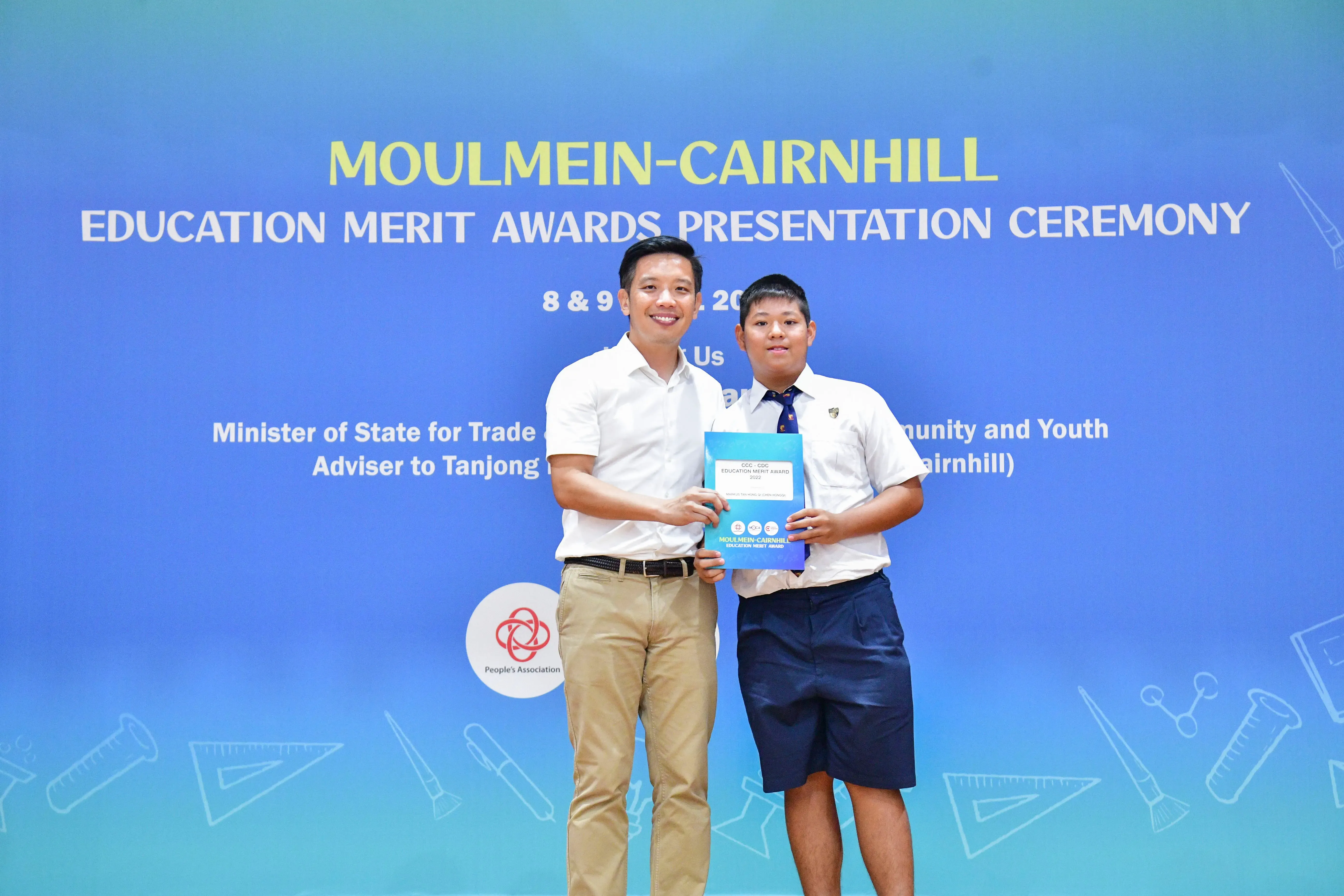 Two people hold an award folder in front of a Moulmein-Cairnhill Education Merit Awards Presentation Ceremony banner.