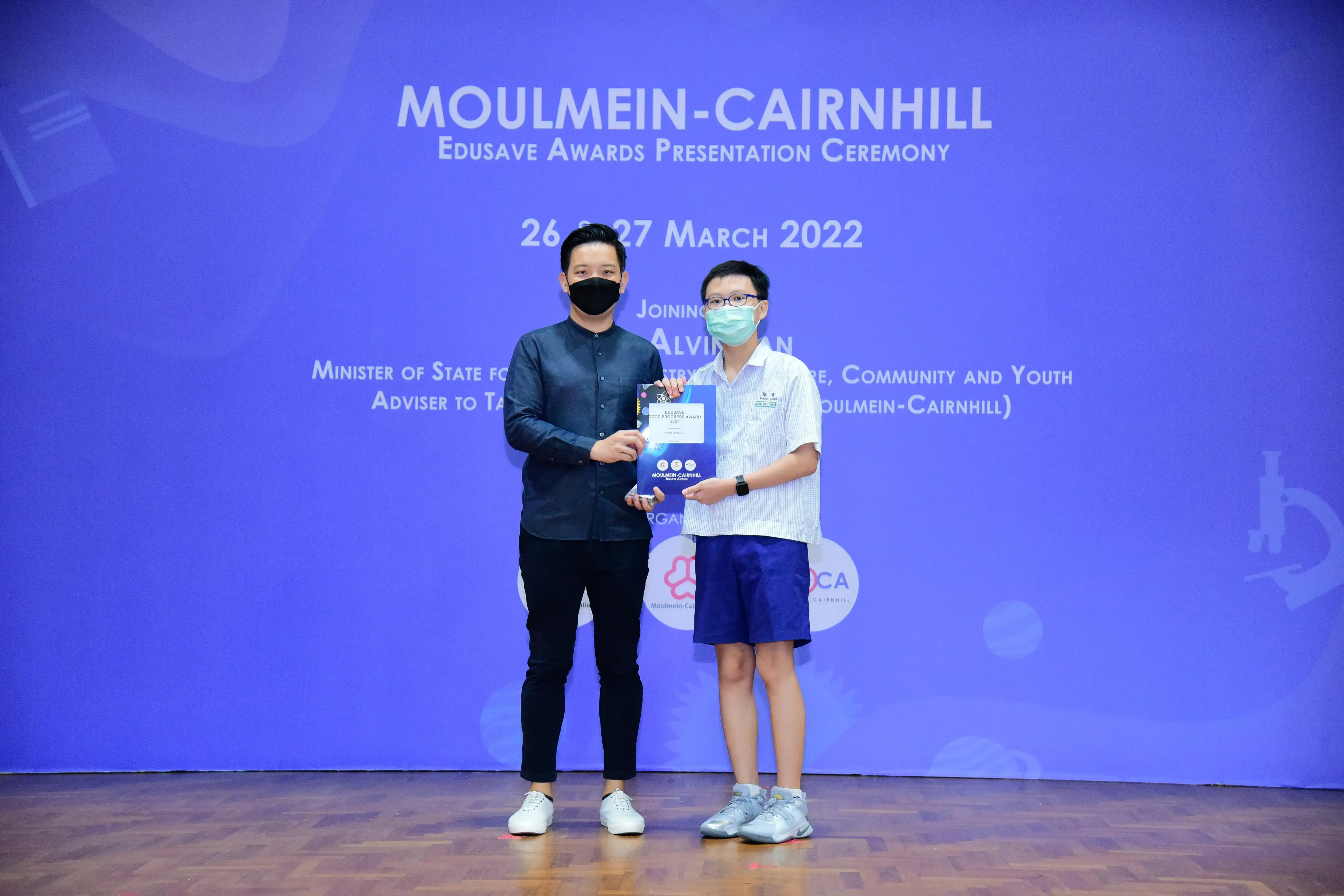 An adult and a young student wearing masks hold an award together at the Moulmein-Cairnhill Edusave Awards Ceremony 2022.