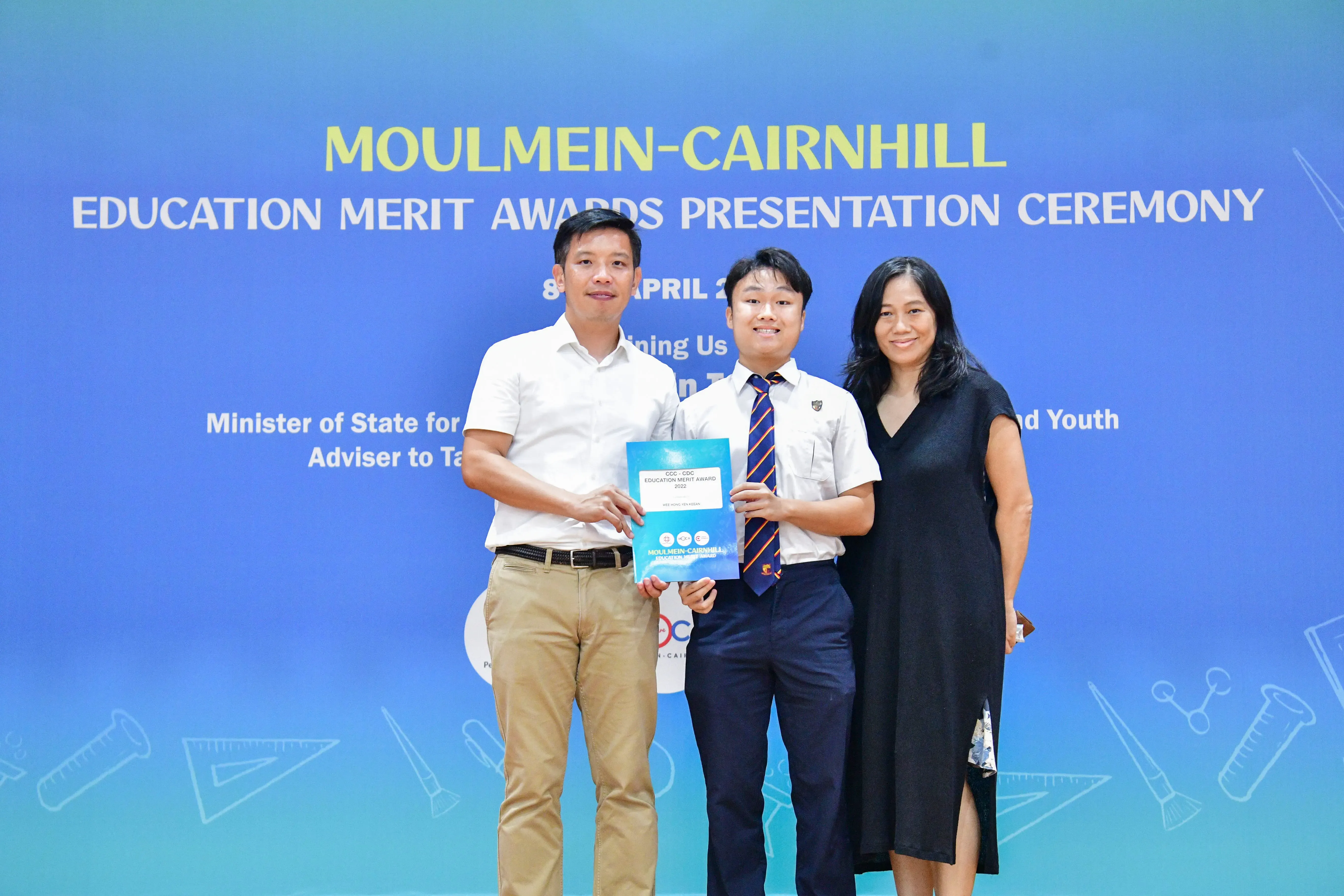Three people pose together at the Moulmein-Cairnhill Education Merit Awards Presentation Ceremony, holding an award booklet.
