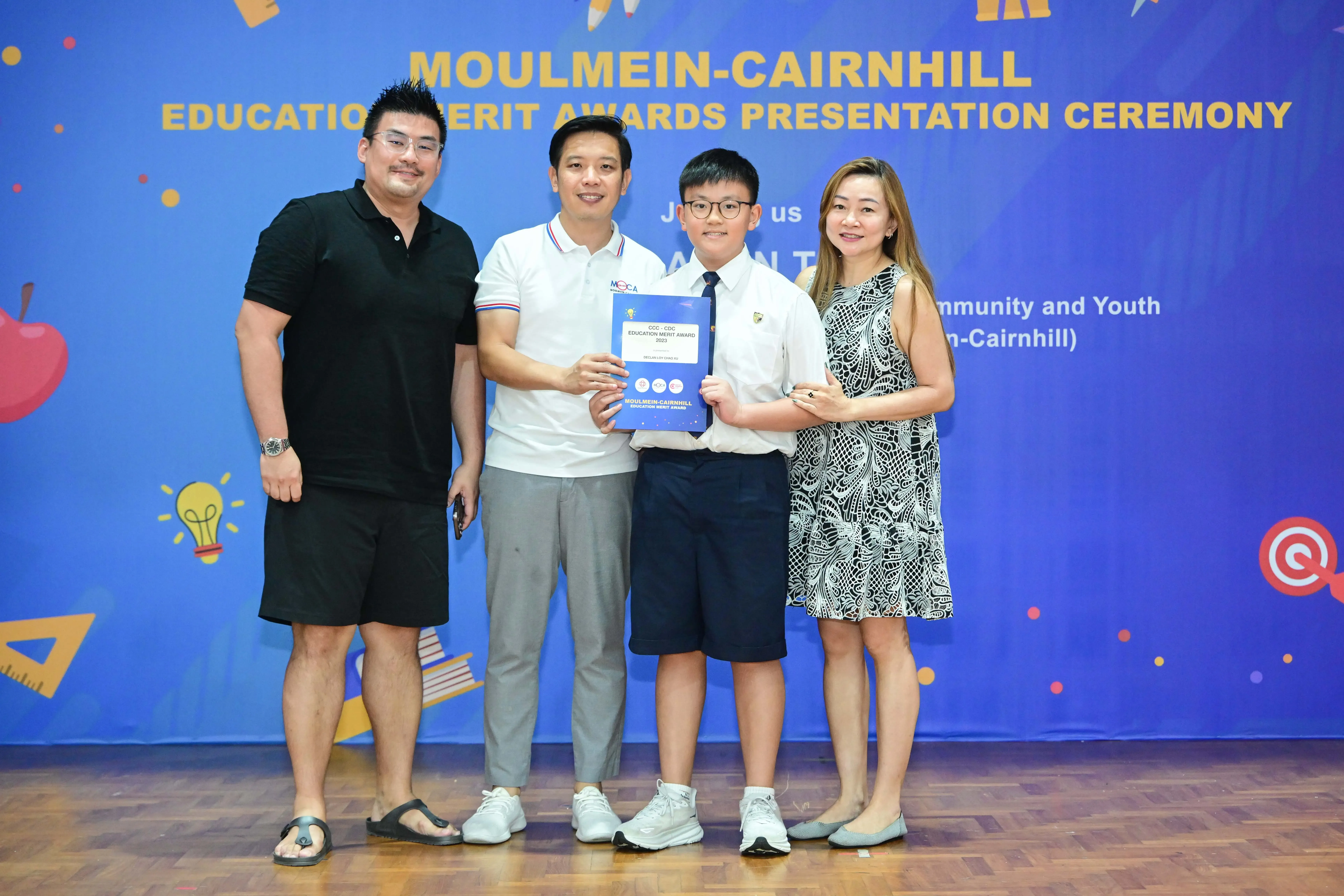 Four people pose at the Moulmein-Cairnhill Education Merit Awards Presentation Ceremony, with a boy holding a certificate.