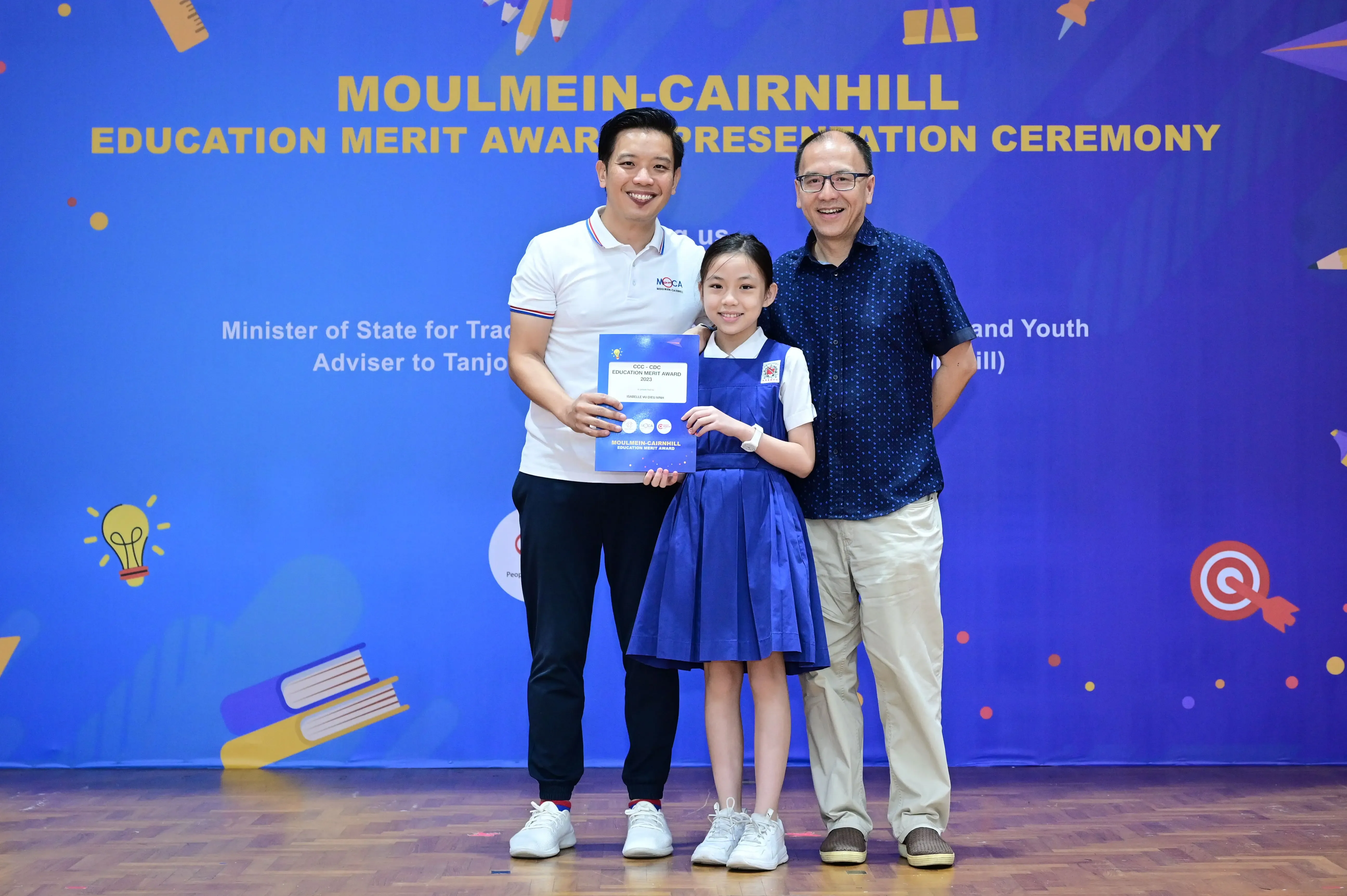 Two men and a girl in a blue uniform hold a certificate at the Moulmein-Cairnhill Education Merit Award ceremony.