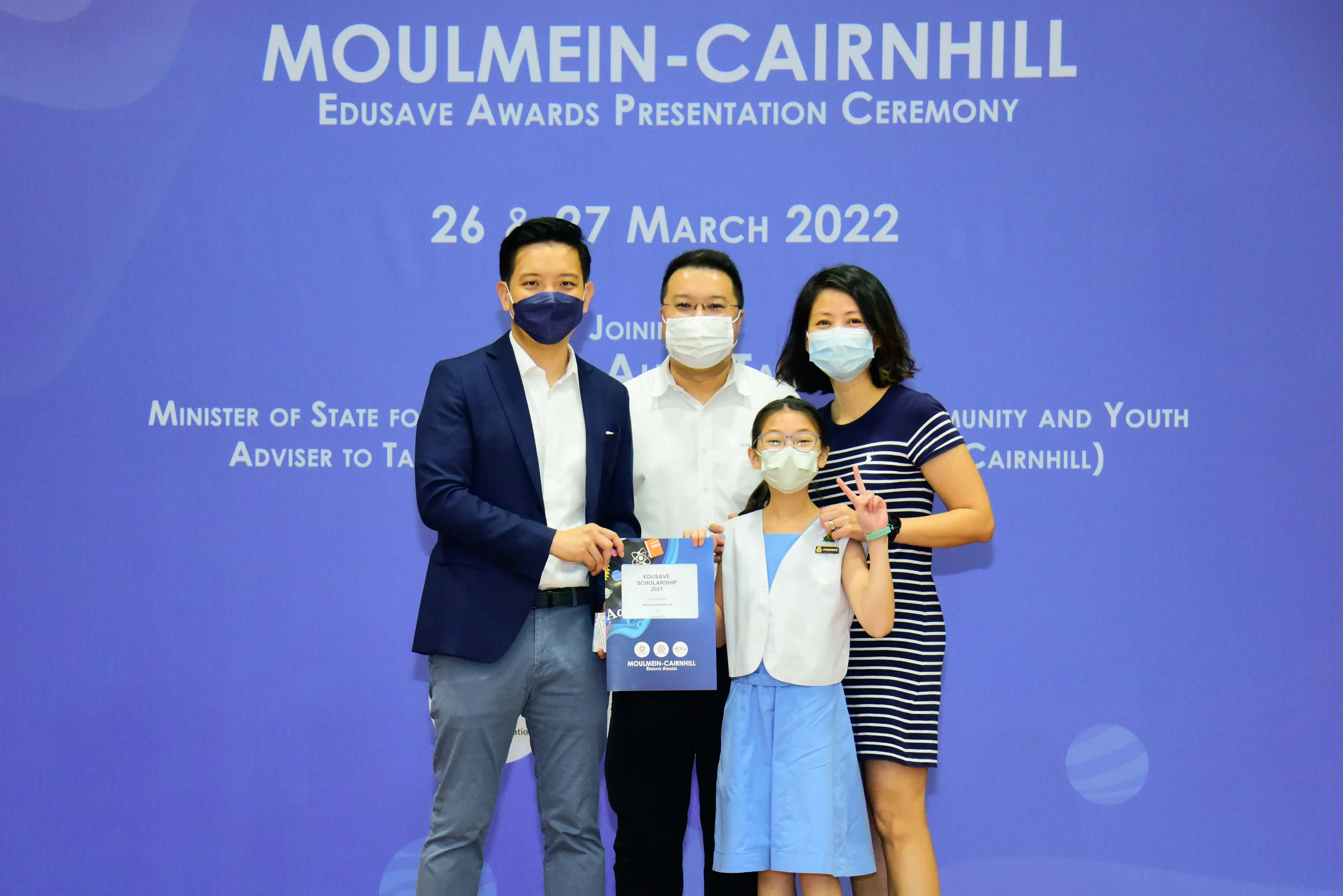 Four masked people pose at the Moulmein-Cairnhill Edusave Awards Ceremony on 26-27 March 2022, with a girl holding a certificate.