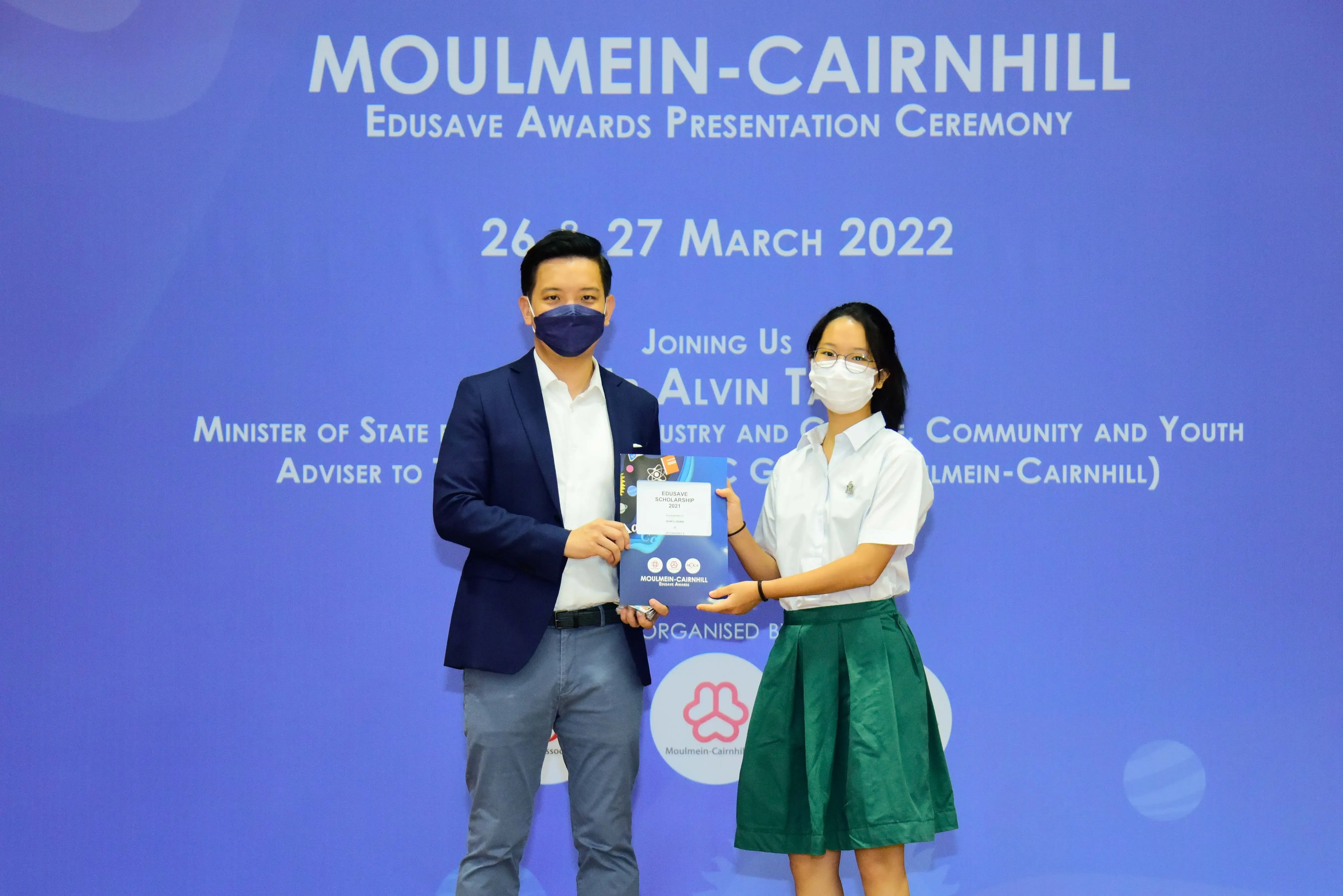 Two masked people hold an Edusave award booklet at the Moulmein-Cairnhill Edusave Awards Ceremony, March 2022.