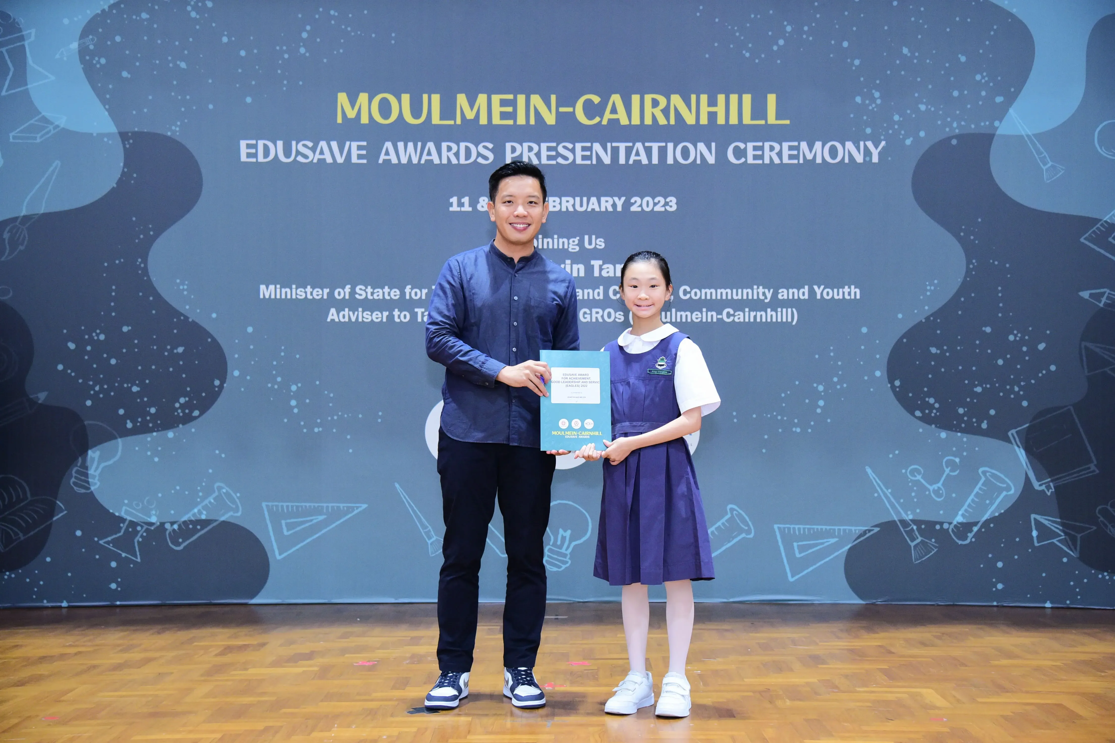 A man and a young girl in school uniform hold a certificate at the Moulmein-Cairnhill Edusave Awards Ceremony 2023.