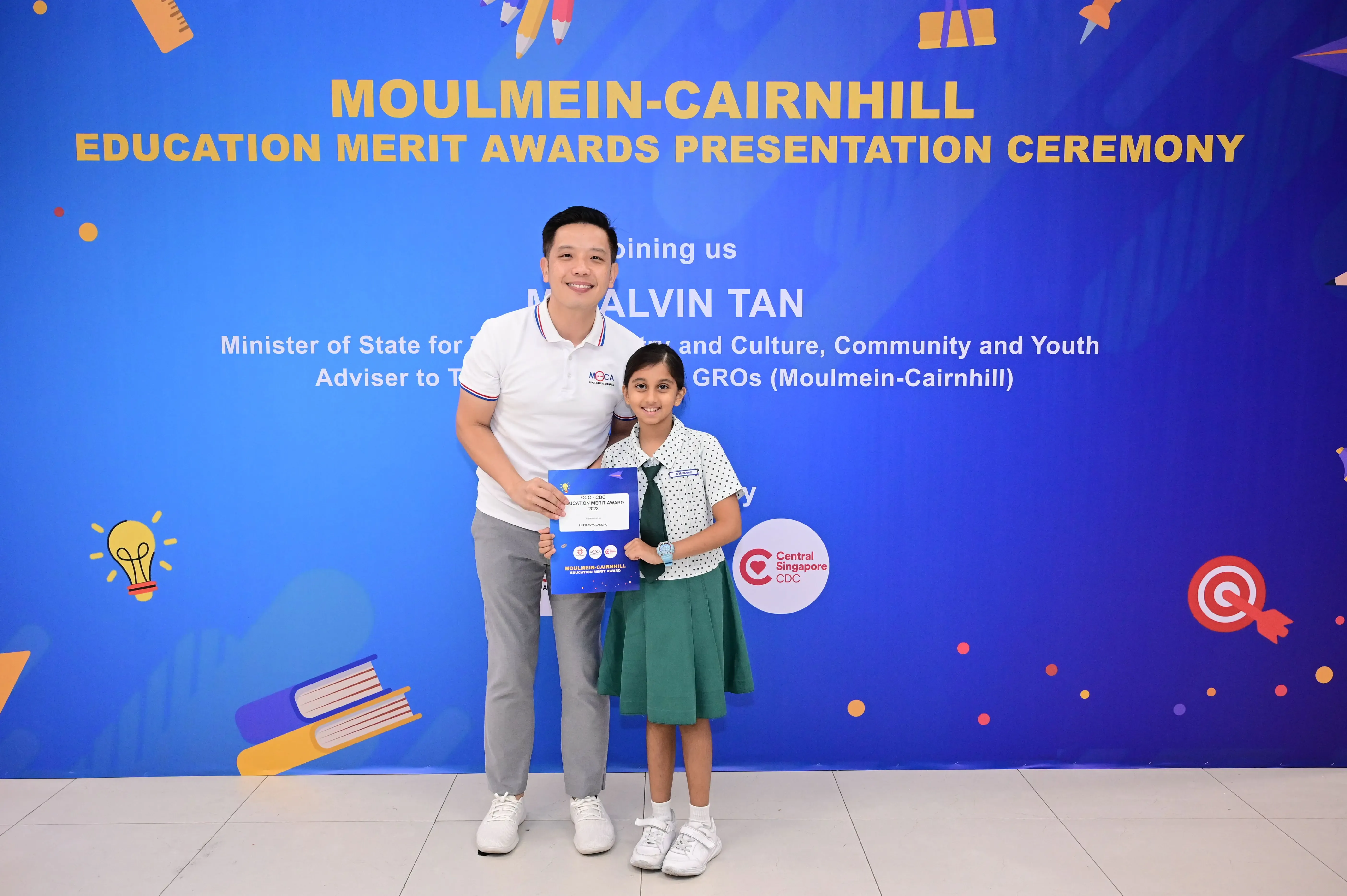A man and a young girl hold an award certificate in front of a Moulmein-Cairnhill Education Merit Awards banner.