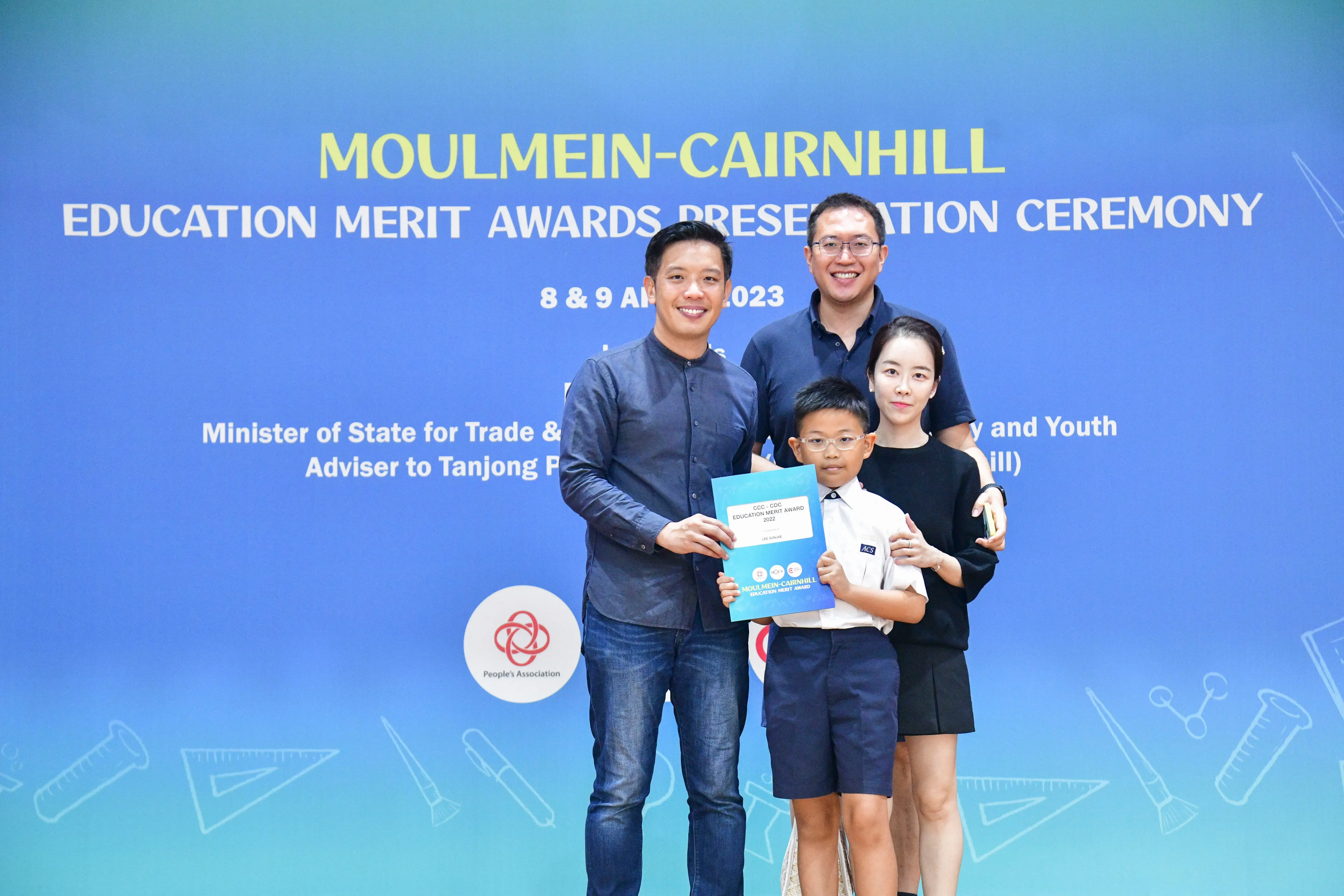 Four people pose before a Moulmein-Cairnhill Education Merit Awards banner; a boy holds a blue certificate.
