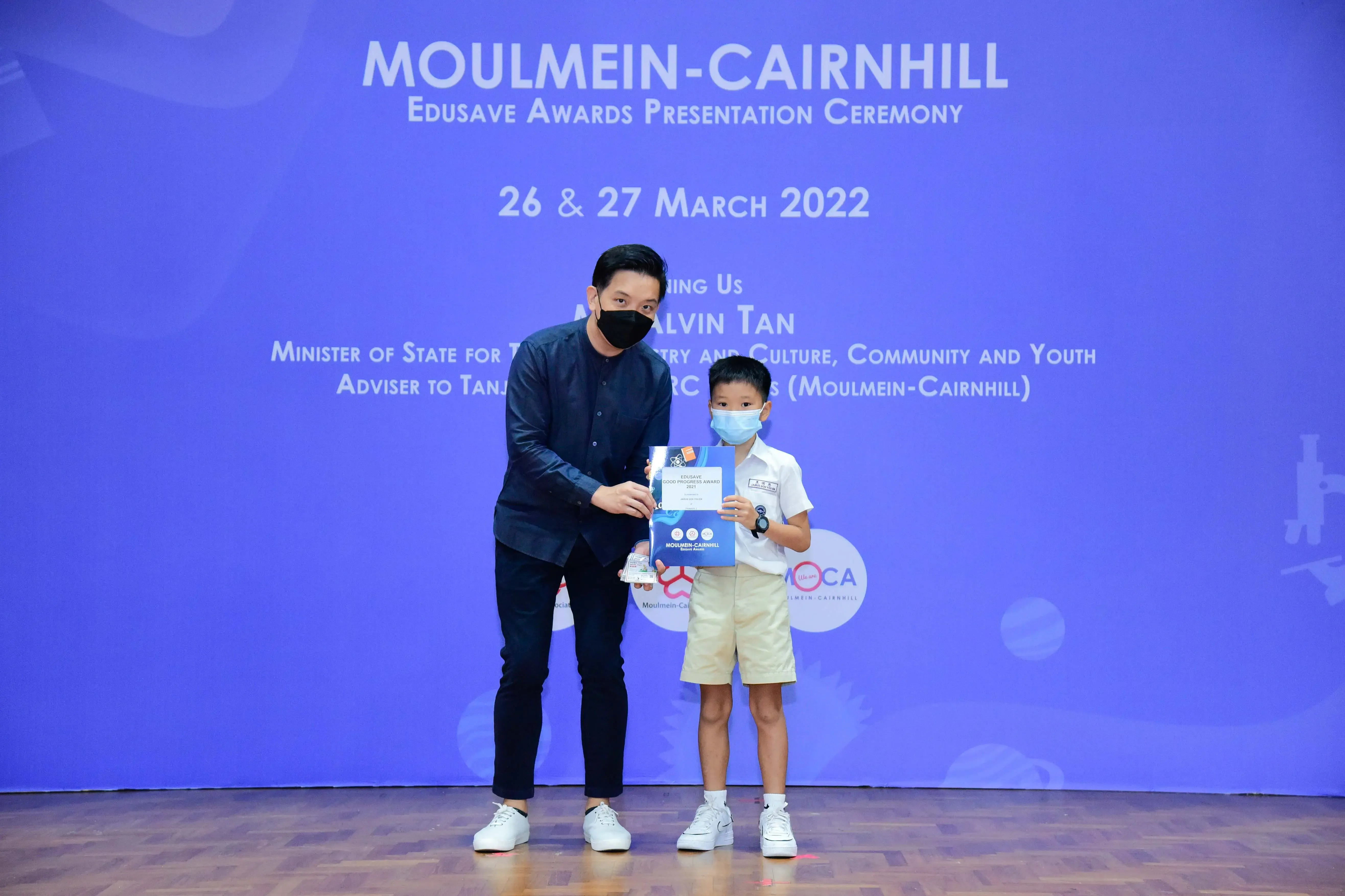 An adult and a young boy in masks hold an award at the Moulmein-Cairnhill Edusave Awards Ceremony, March 2022.