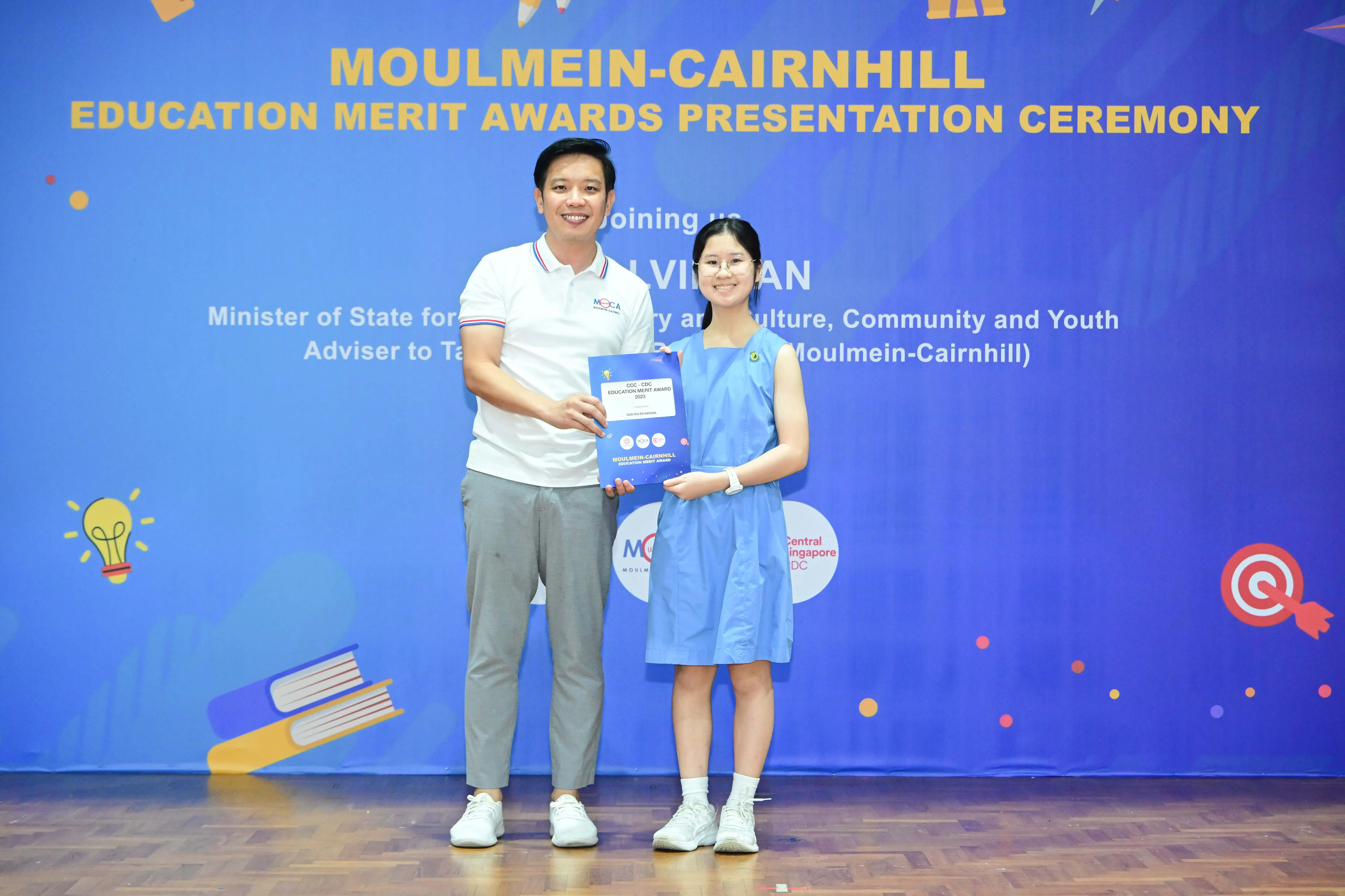A man and a young girl hold a certificate together at the Moulmein-Cairnhill Education Merit Awards Presentation Ceremony.